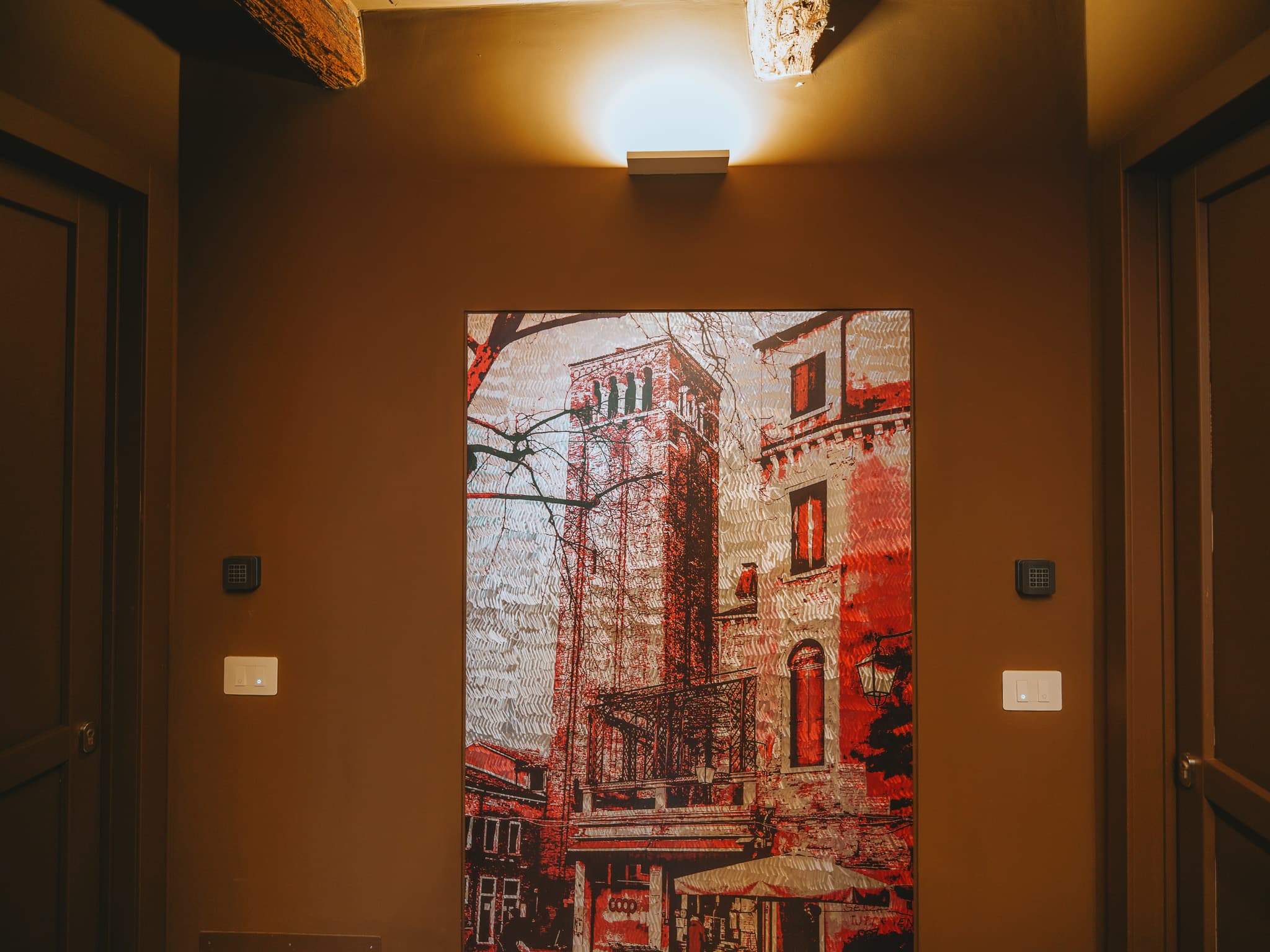 Stampa incorniciata in toni di rosso con un campanile e case veneziane, illuminata da una lampada a parete, tra due porte scure.