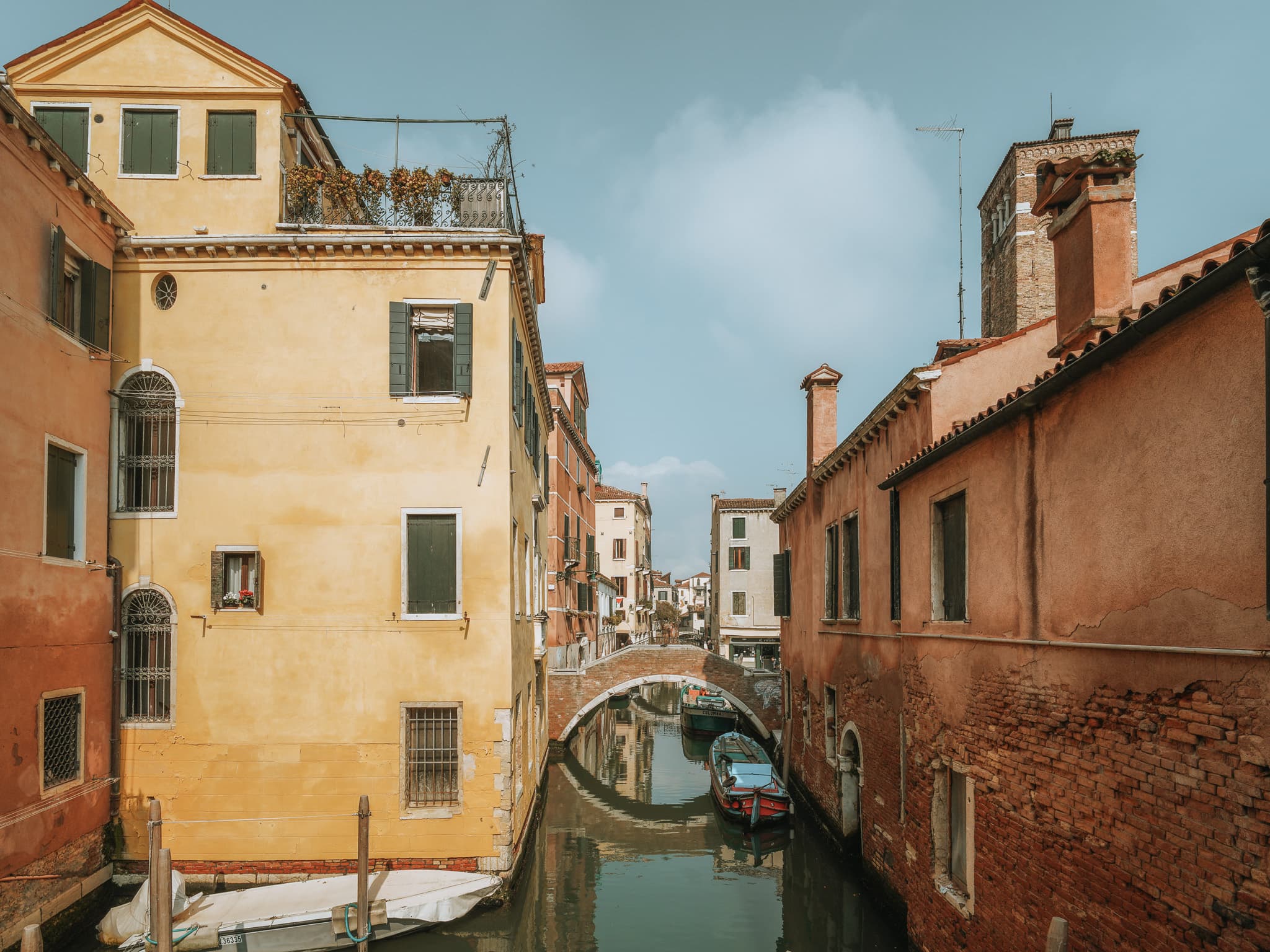 Canale stretto con barche ormeggiate, palazzo ocra con persiane grigio-verdi e, sullo sfondo, un ponte ad arco in mattoni e la sommita' di un campanile.