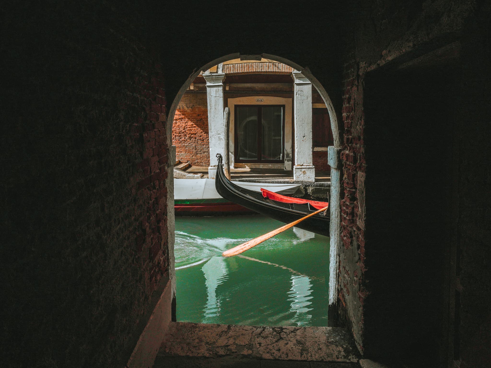 Vista da un sottoportego in mattoni su un canale verde: l'estremita' nera ricurva di una gondola, un remo chiaro appoggiato sull'acqua e la facciata opposta con colonne chiare.