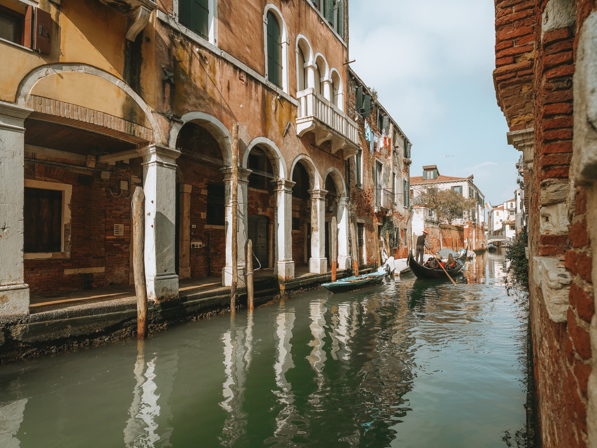 Canale con portico ad archi, pali di ormeggio e un gondoliere che voga con dei passeggeri.