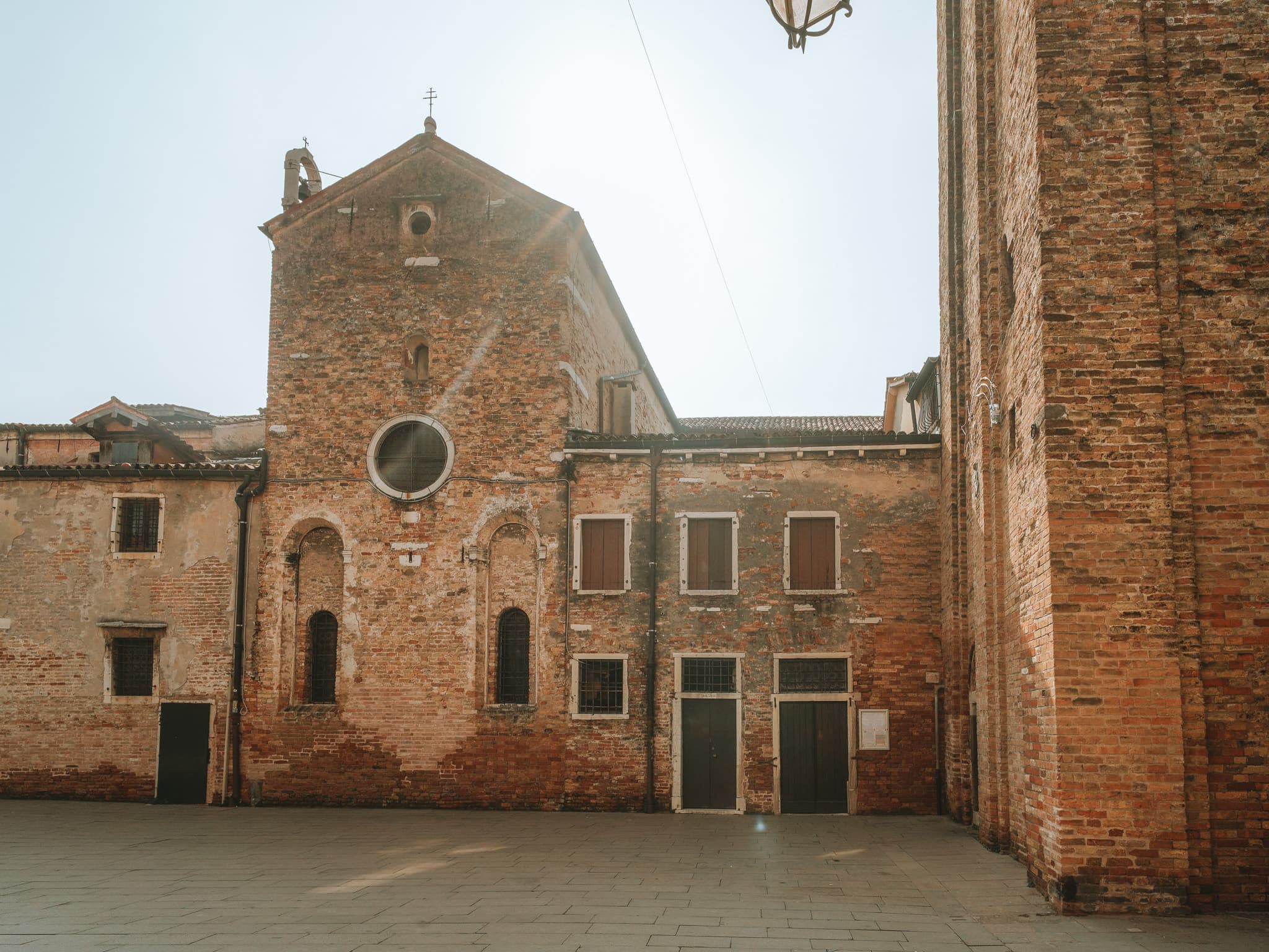 Facciata di chiesa in mattoni con finestra circolare, due finestre ad arco con grate e campaniletto a vela; a destra la base in mattoni del campanile.