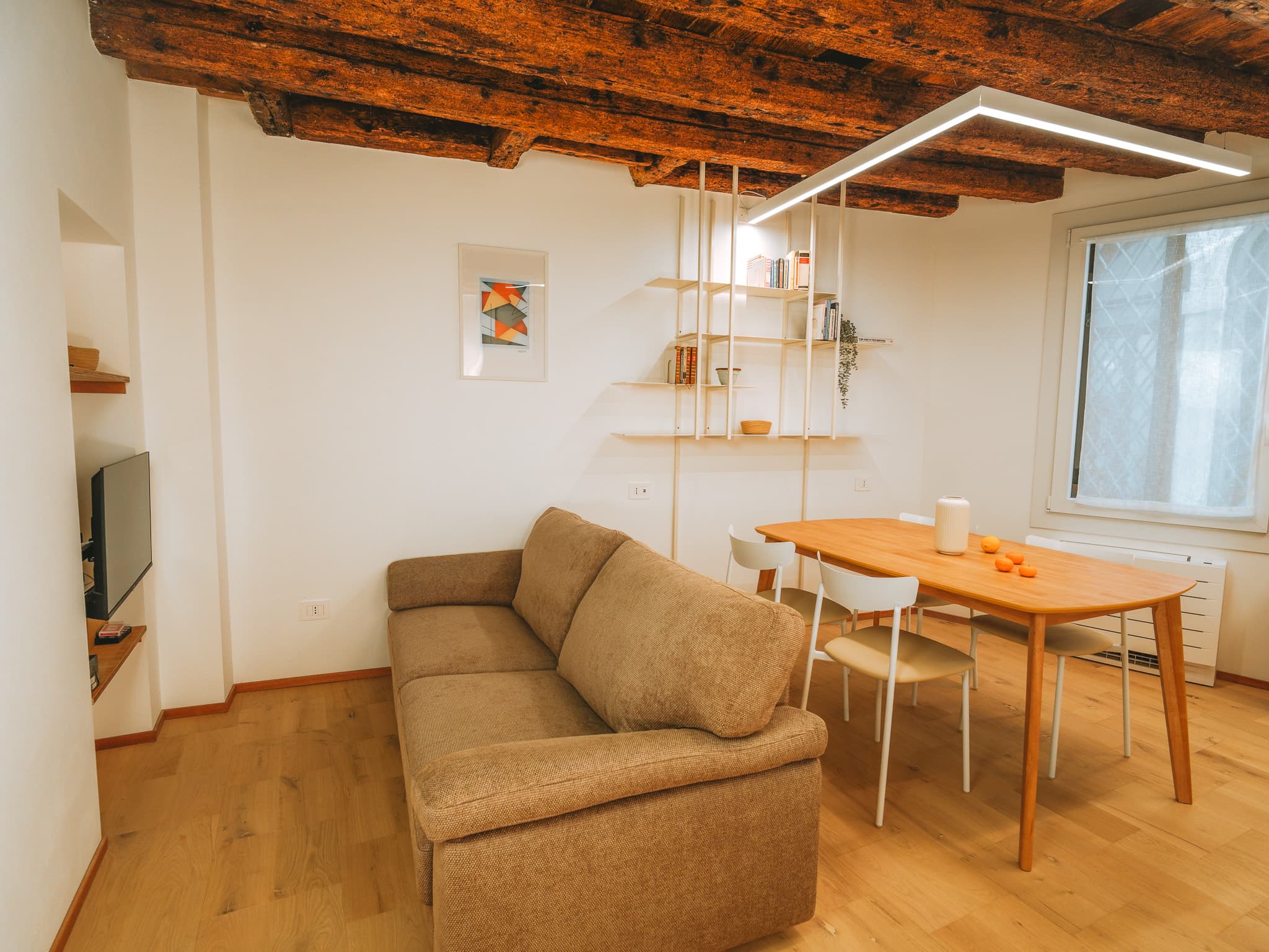 Soggiorno con divano beige, tavolo da pranzo in legno chiaro con sedie bianche, libreria bianca a parete e soffitto con travi di legno a vista