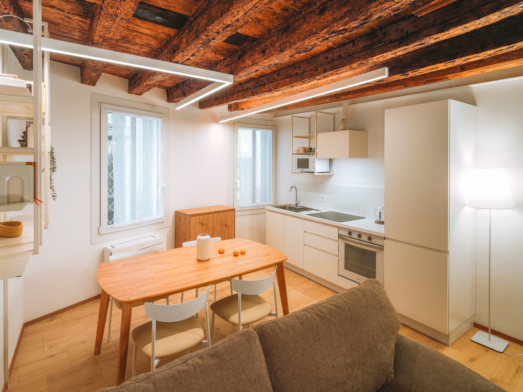 Cucina lineare bianca con lavello, piano cottura nero, forno e colonna frigo, tavolo in legno con sedie bianche e due finestre