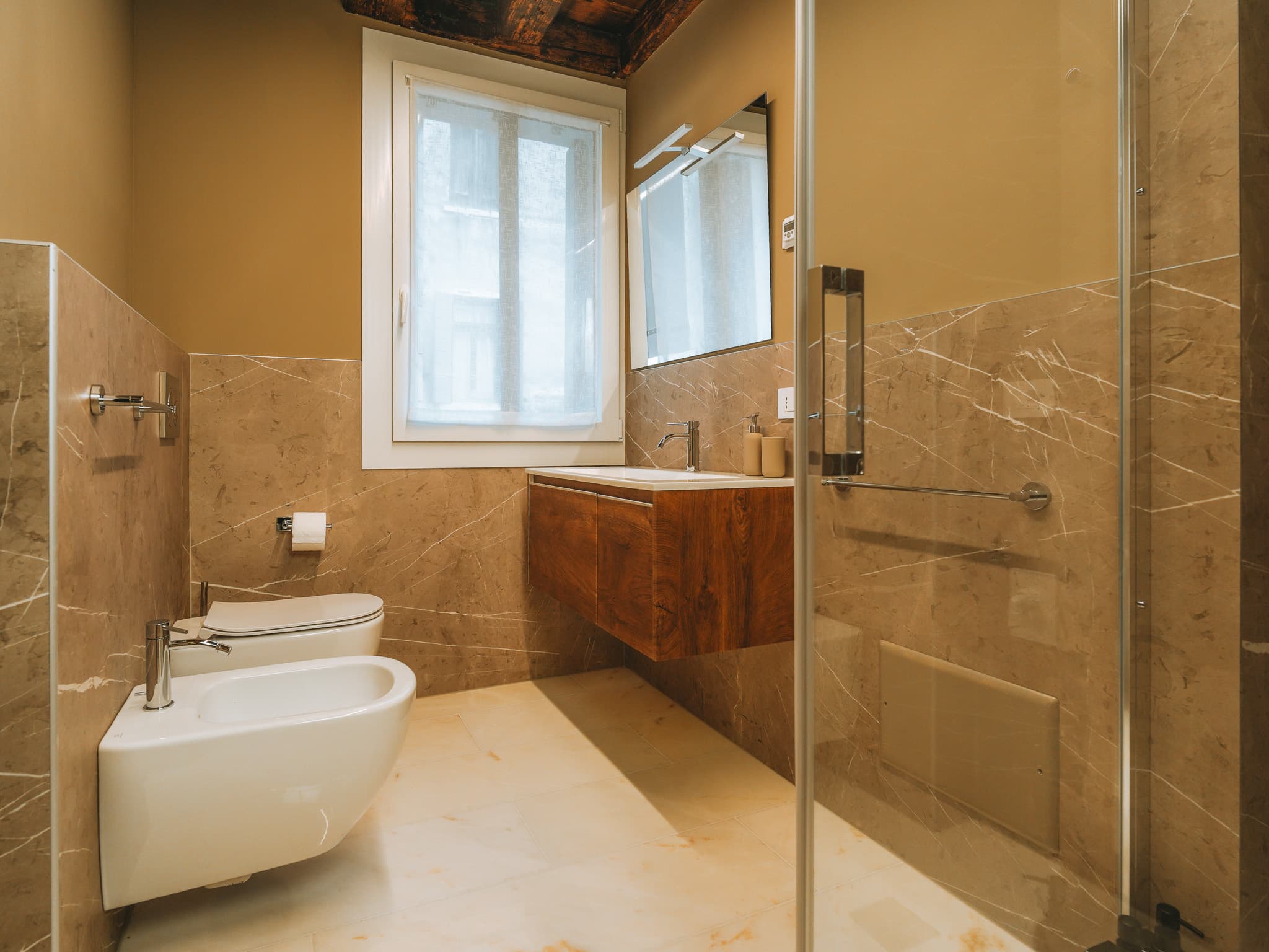 Bagno con pareti in marmo beige a mezza altezza, wc e bidet sospesi, mobile lavabo in legno scuro con specchio, finestra e box doccia in vetro