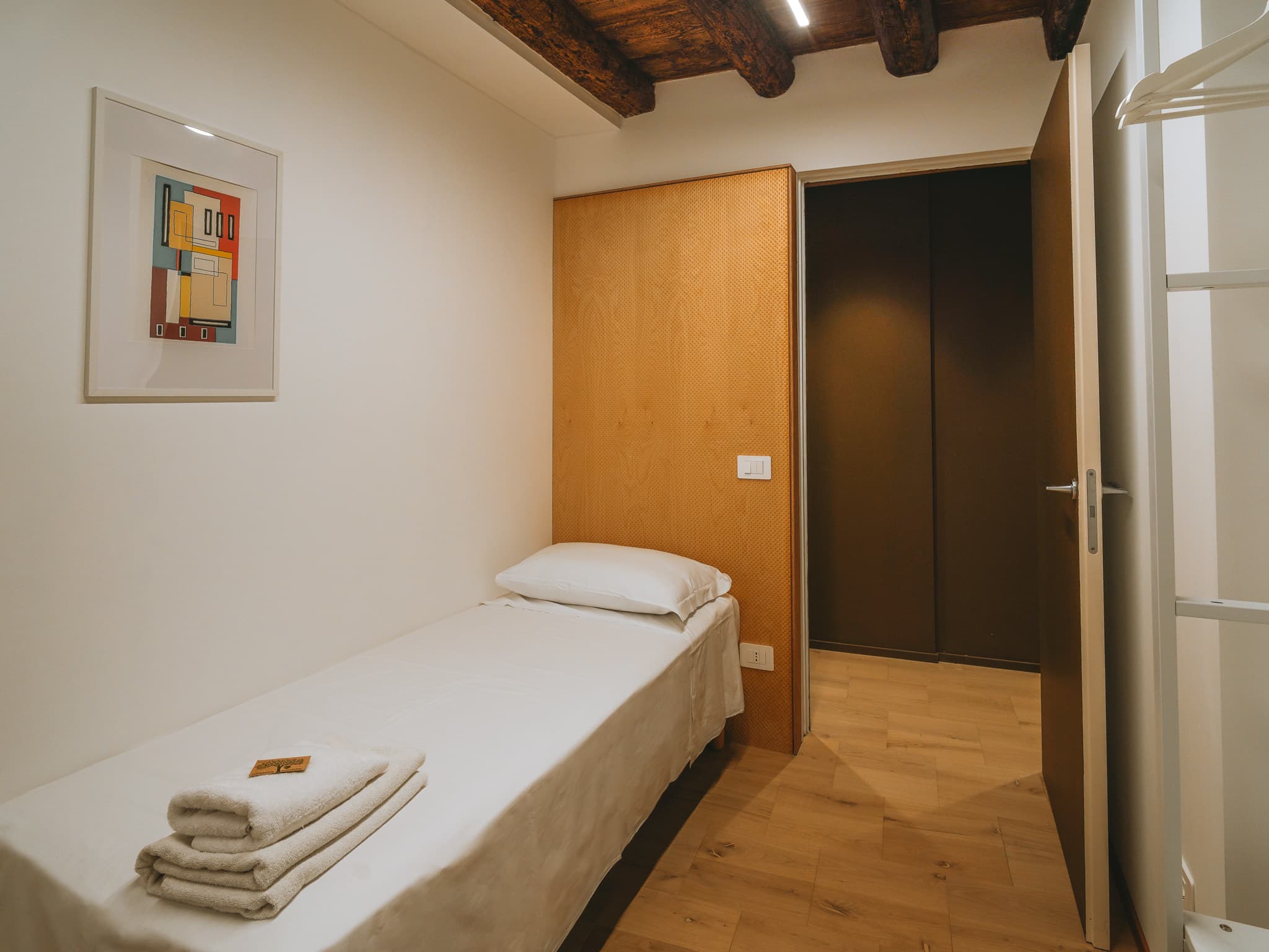Letto singolo con biancheria bianca e asciugamani piegati, pannello a parete in legno chiaro a trama intrecciata, stampa geometrica incorniciata e porta bianca aperta