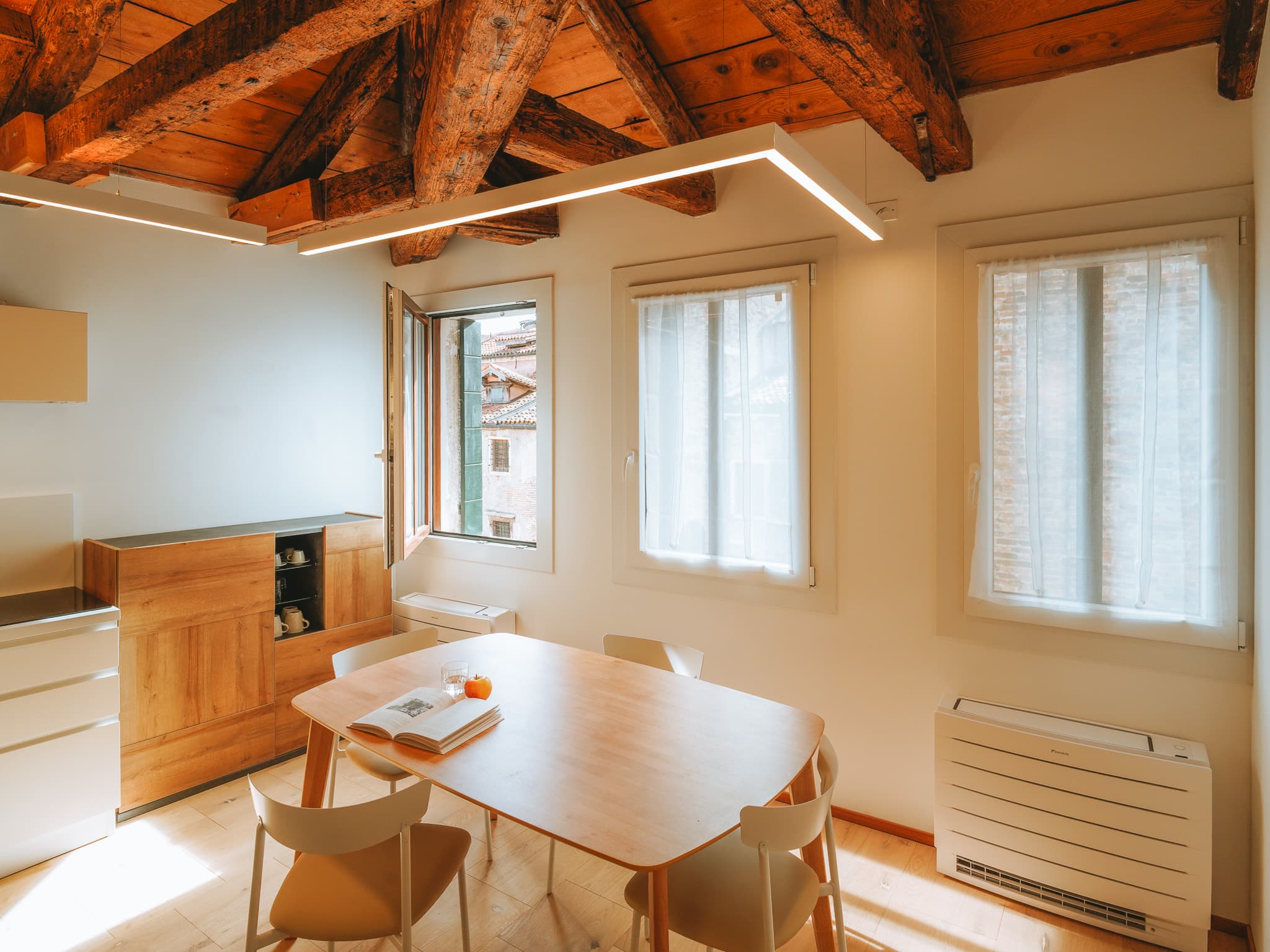 Tavolo ovale in legno con quattro sedie chiare, finestra aperta sui tetti e soffitto a travi