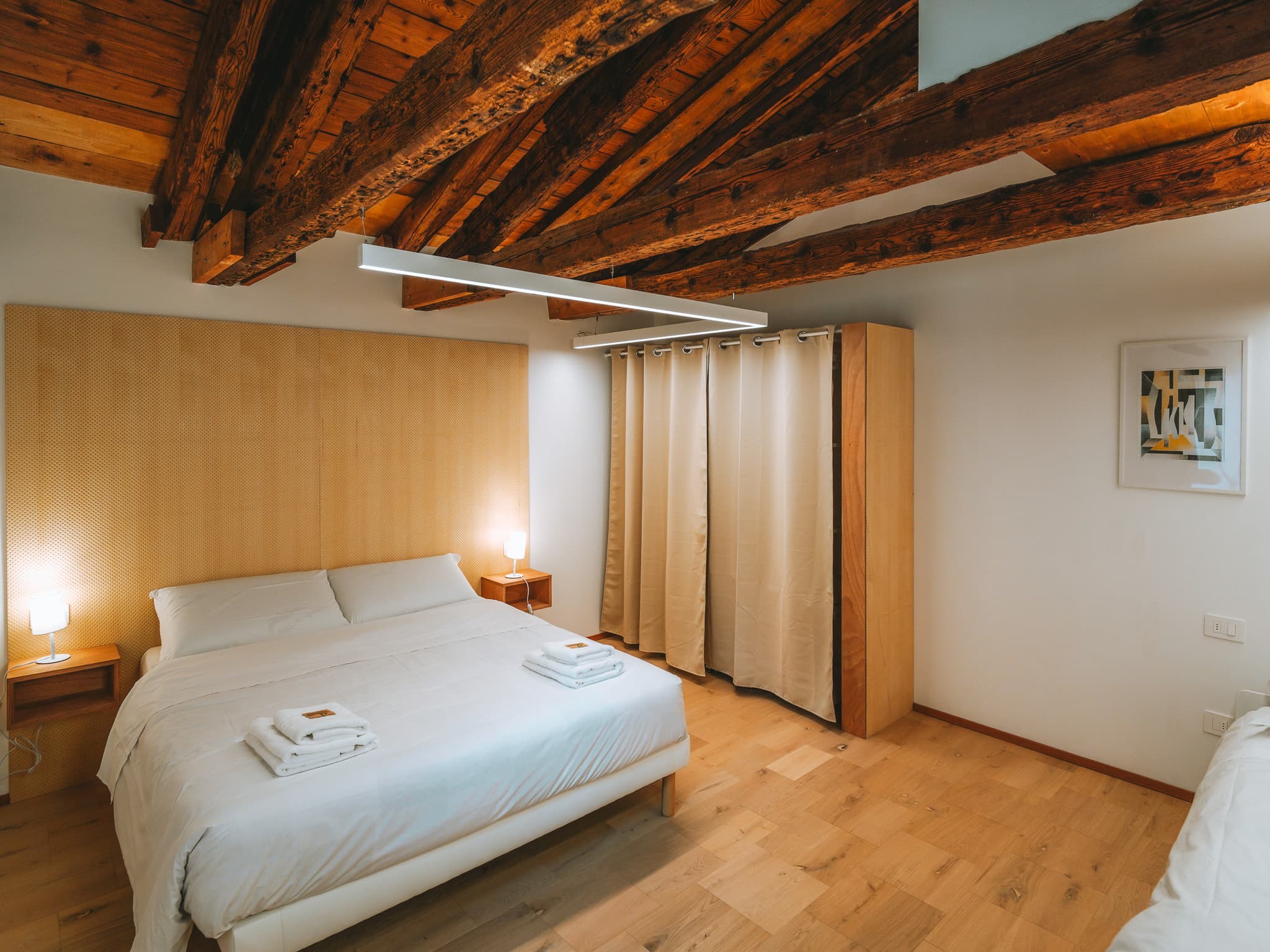 Letto matrimoniale con testiera a tutta parete, due comodini con lampade accese e travi a vista