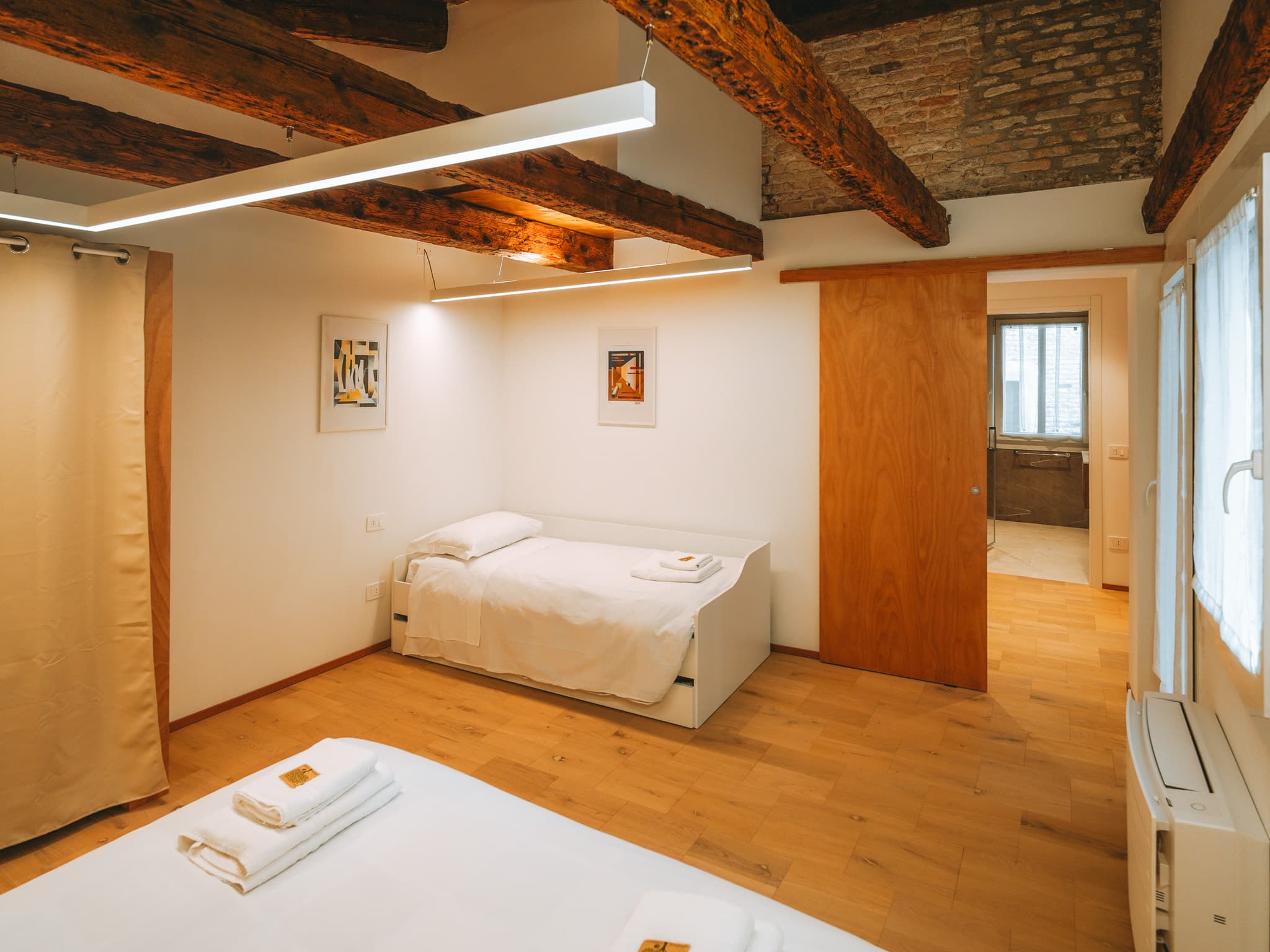 Letto singolo bianco, porta scorrevole in legno aperta sul bagno e soffitto con travi e mattoni