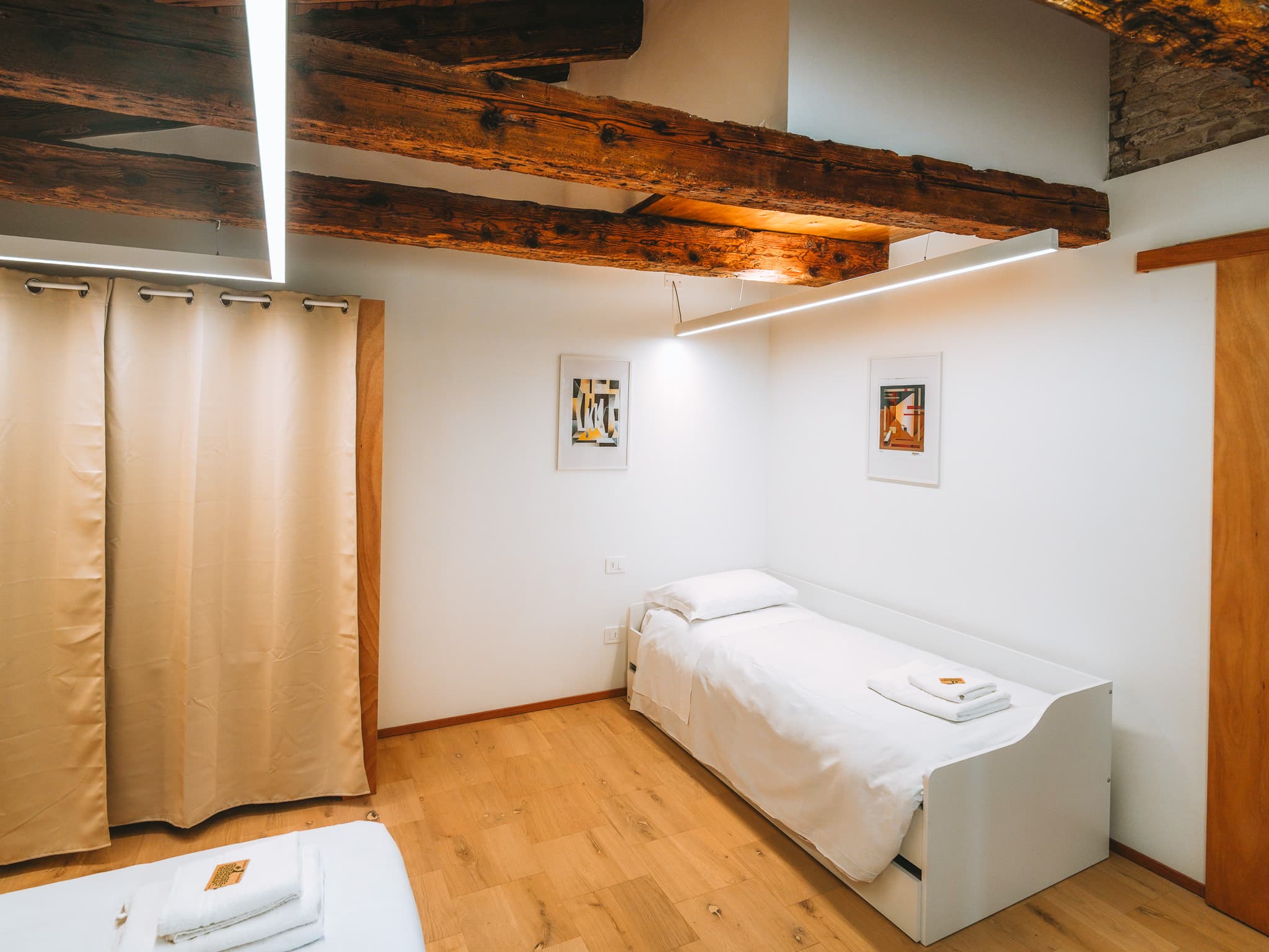 Letto singolo bianco contro la parete con due stampe incorniciate e tenda beige a lato