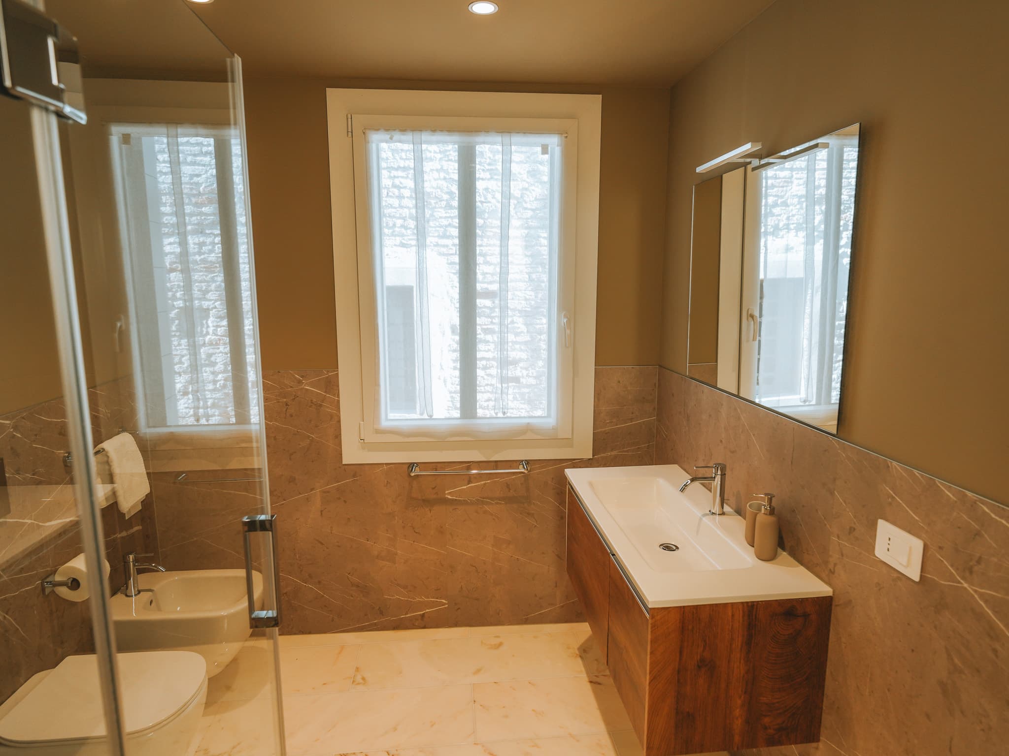 Bagno con marmo beige, lavabo bianco su mobile in legno, bidet e box doccia in vetro.