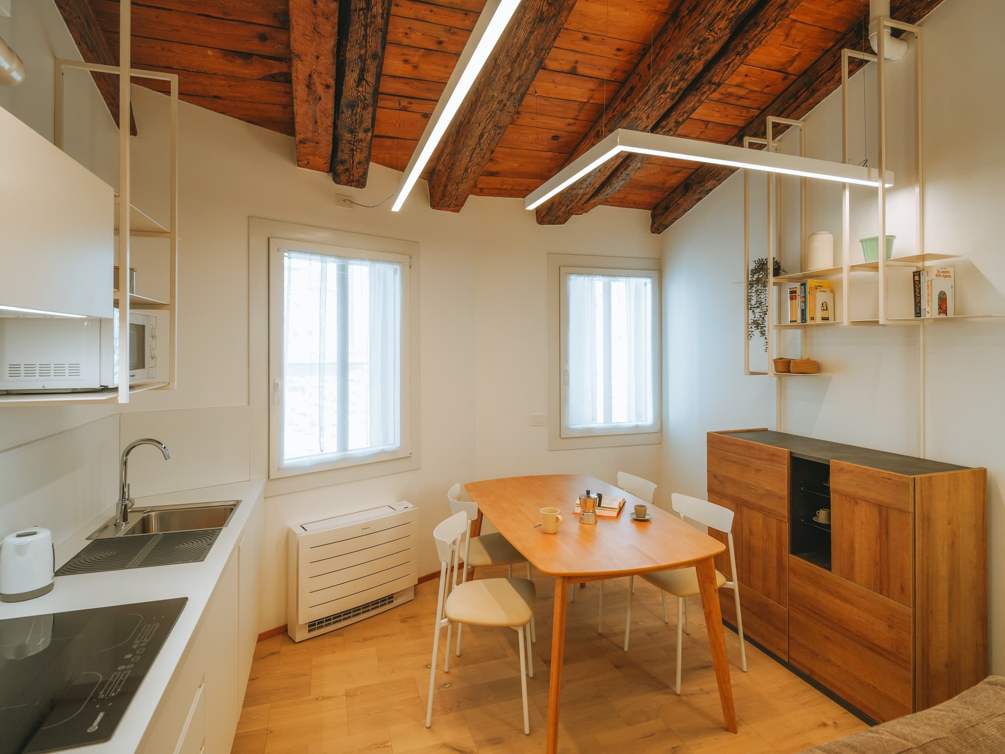 Cucina bianca lineare con tavolo ovale in legno, sedie bianche e soffitto con travi a vista.