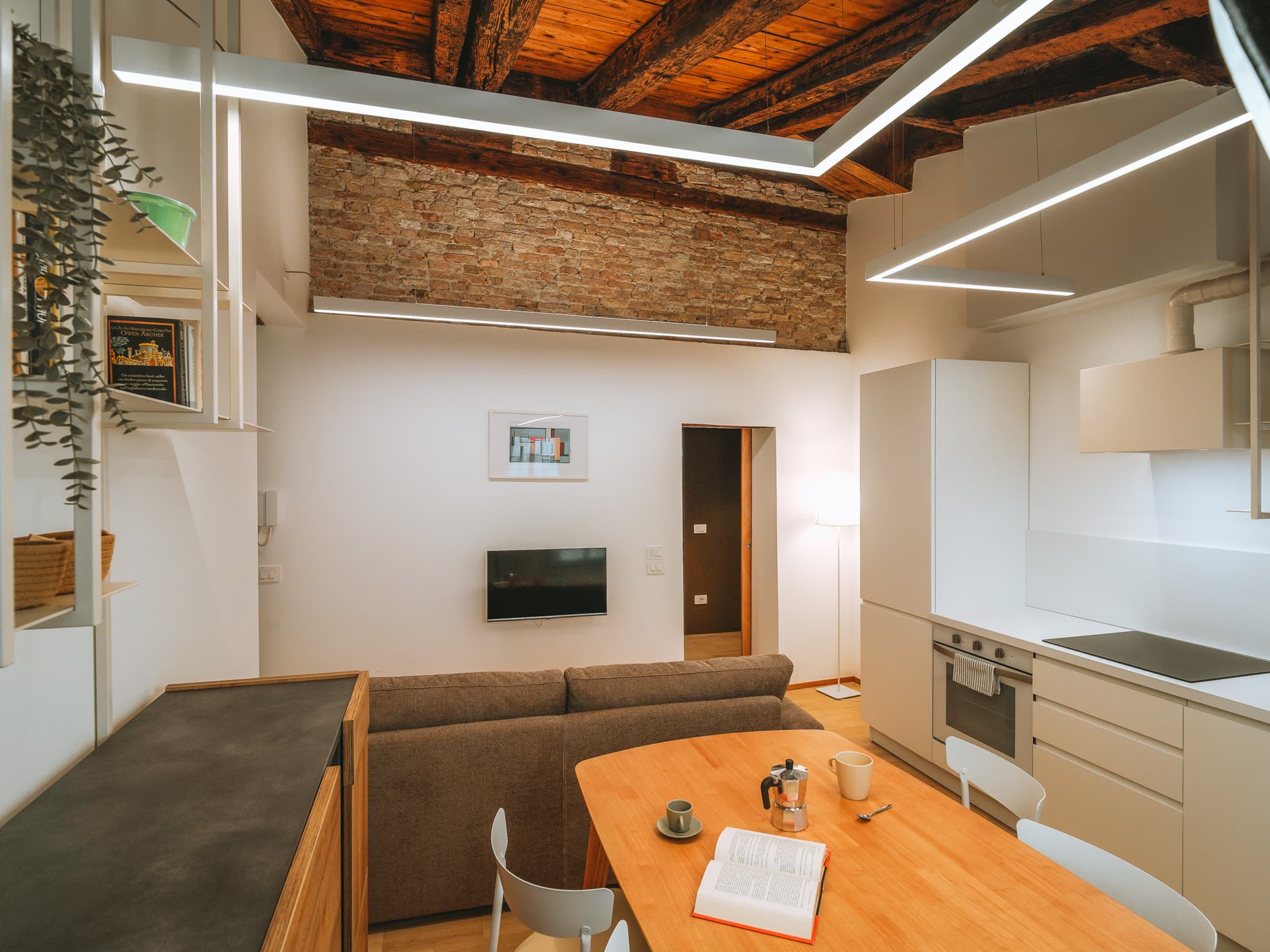 Zona giorno con divano, televisore a parete e tavolo in legno; fascia di mattoni a vista sotto il soffitto travato e cucina bianca sulla destra.