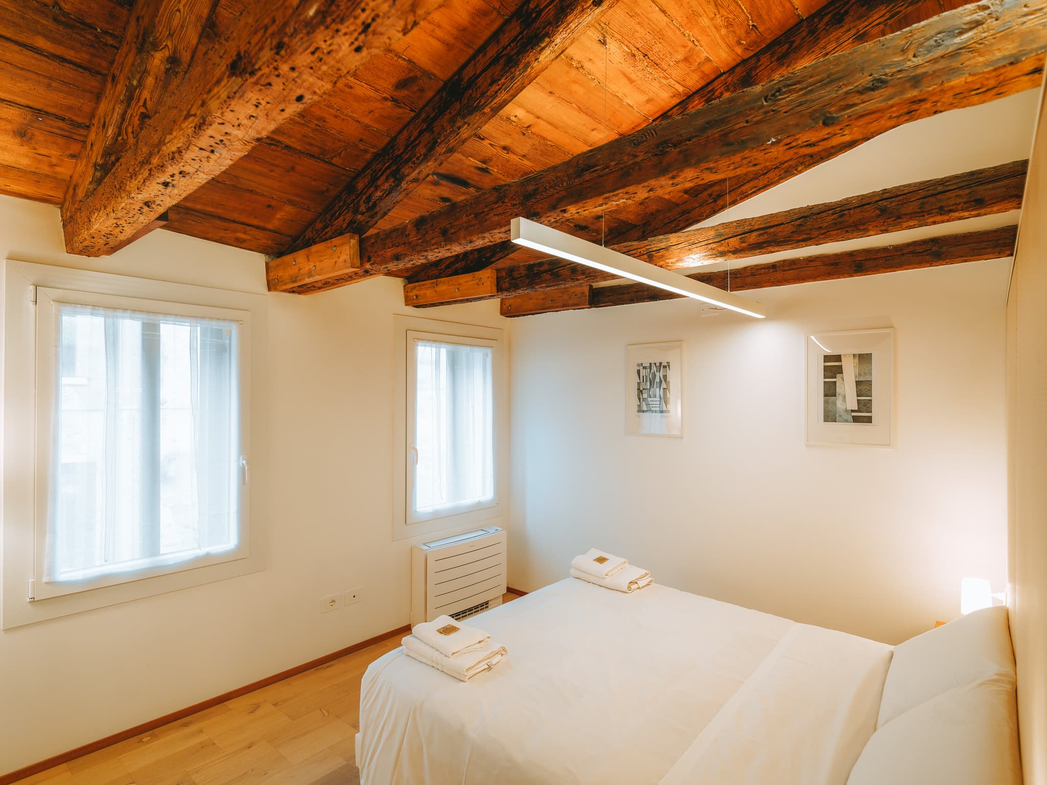 Camera con letto bianco, due finestre con tende chiare, travi a vista e due stampe incorniciate.