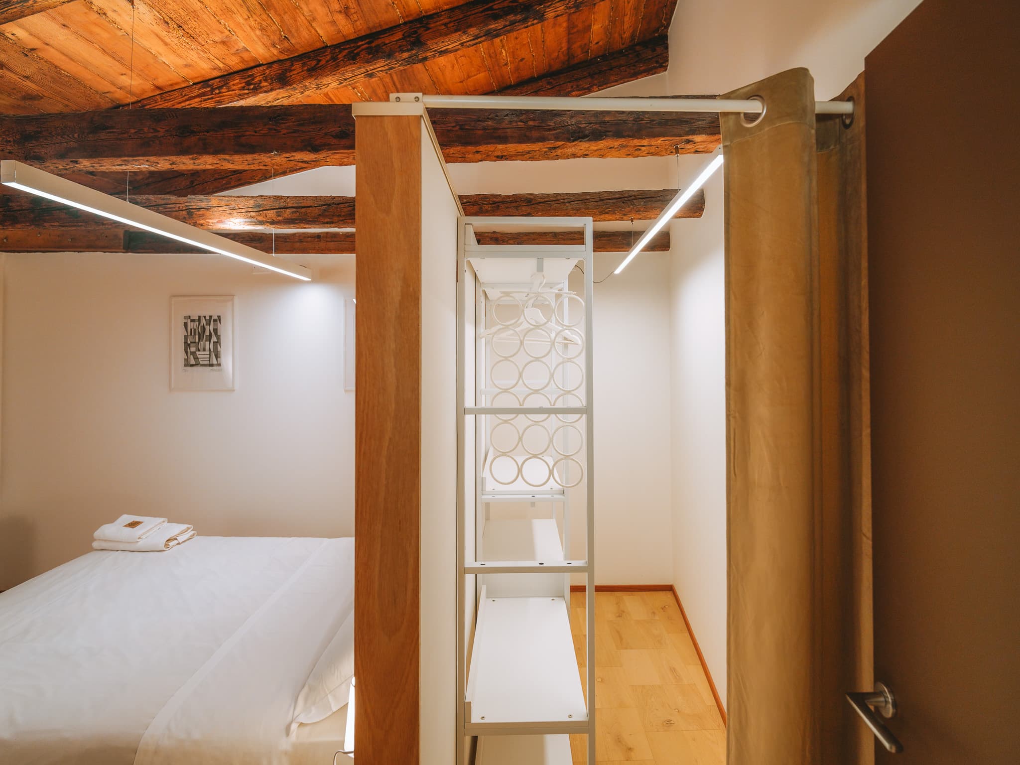Armadio aperto con ripiani bianchi e grucce dietro una tenda beige; a sinistra il letto bianco con asciugamani piegati.