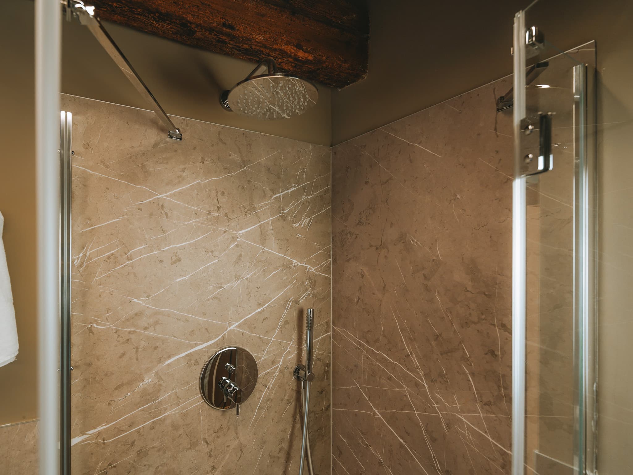 Doccia rivestita in marmo beige venato, soffione a pioggia cromato e trave di legno sopra.