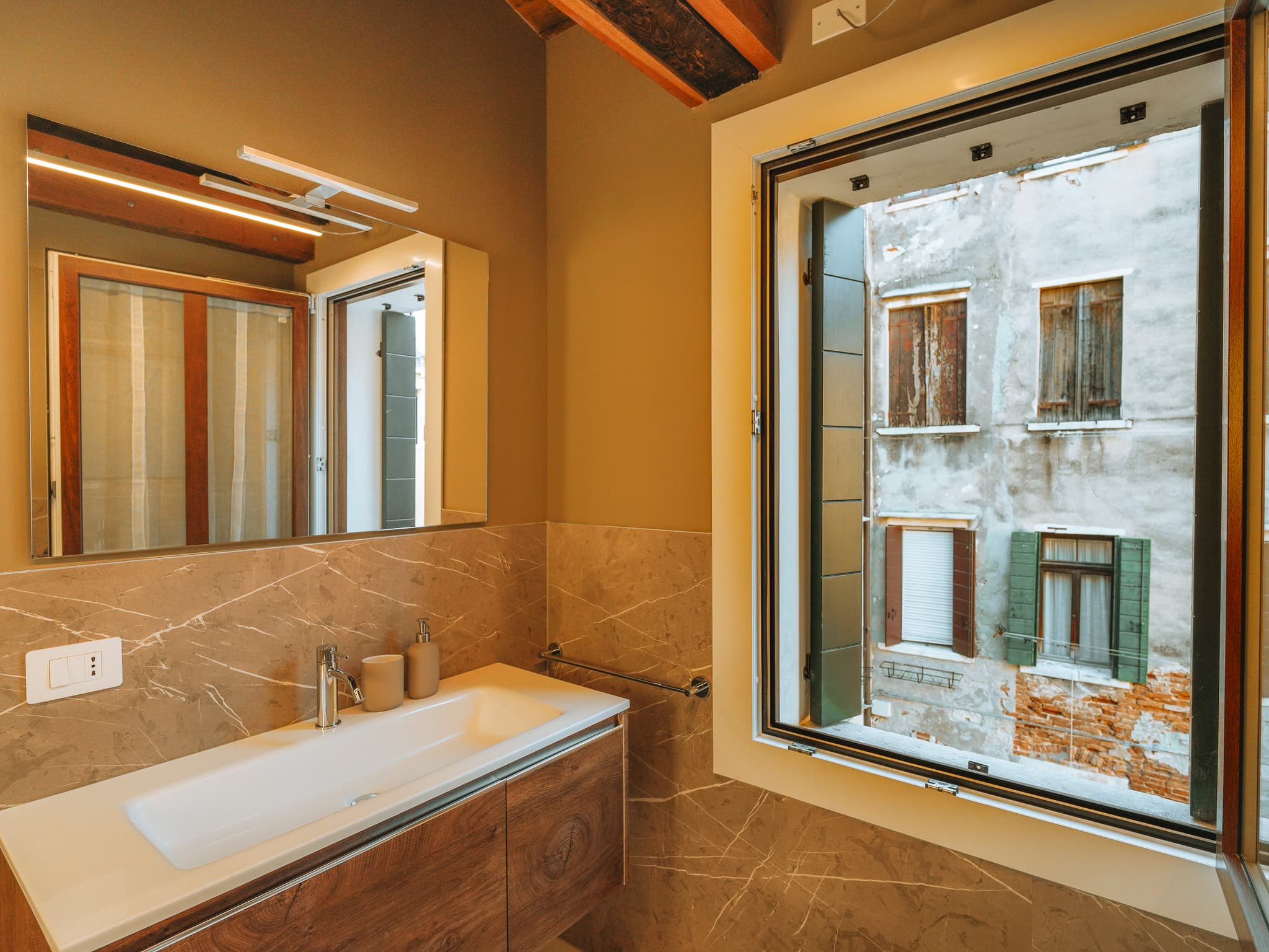 Bagno con lavabo bianco, specchio e finestra aperta su una facciata con persiane e intonaco scrostato.