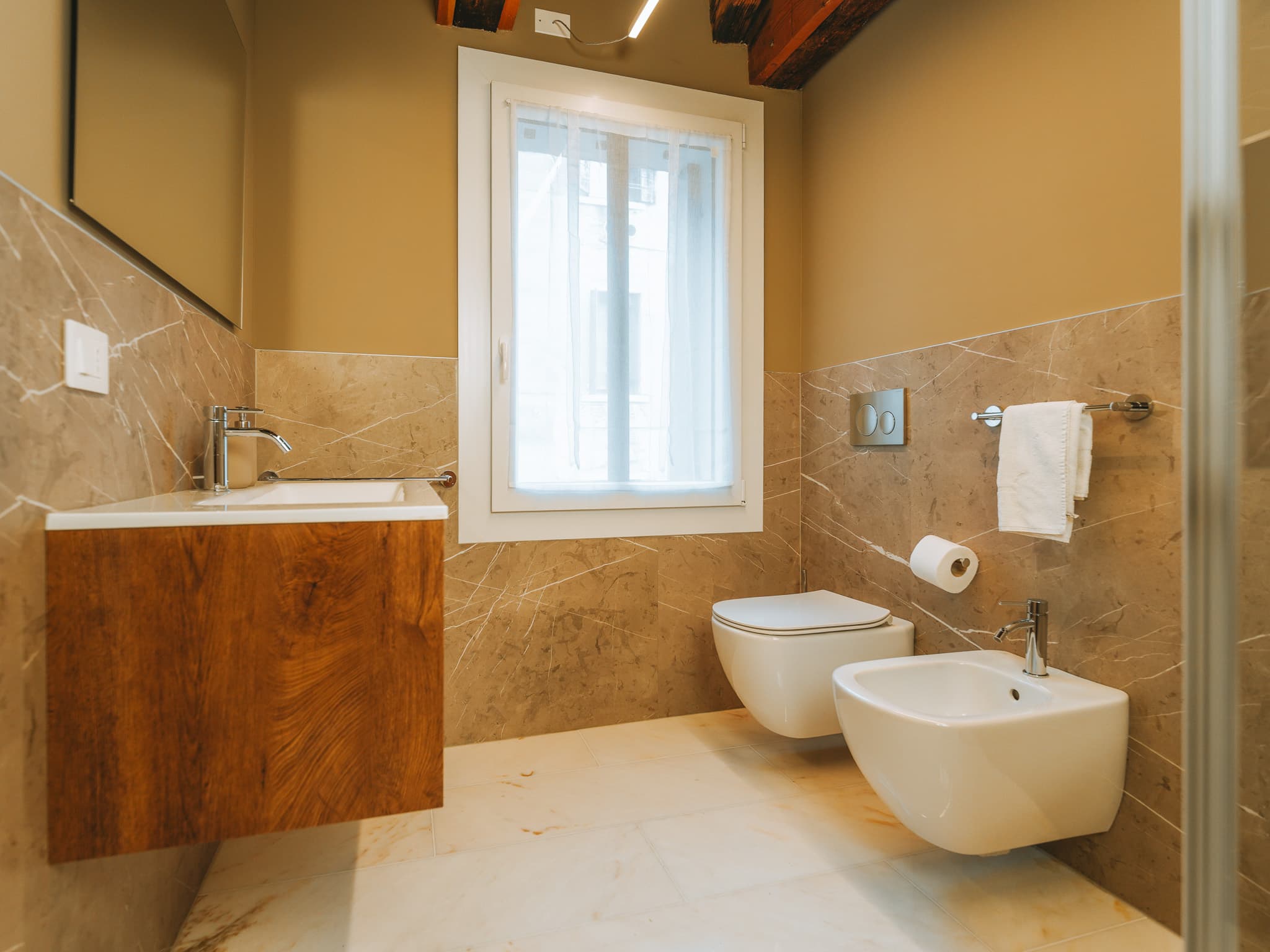 Bagno con lavabo su mobile in legno, wc e bidet sospesi, finestra con tenda bianca.