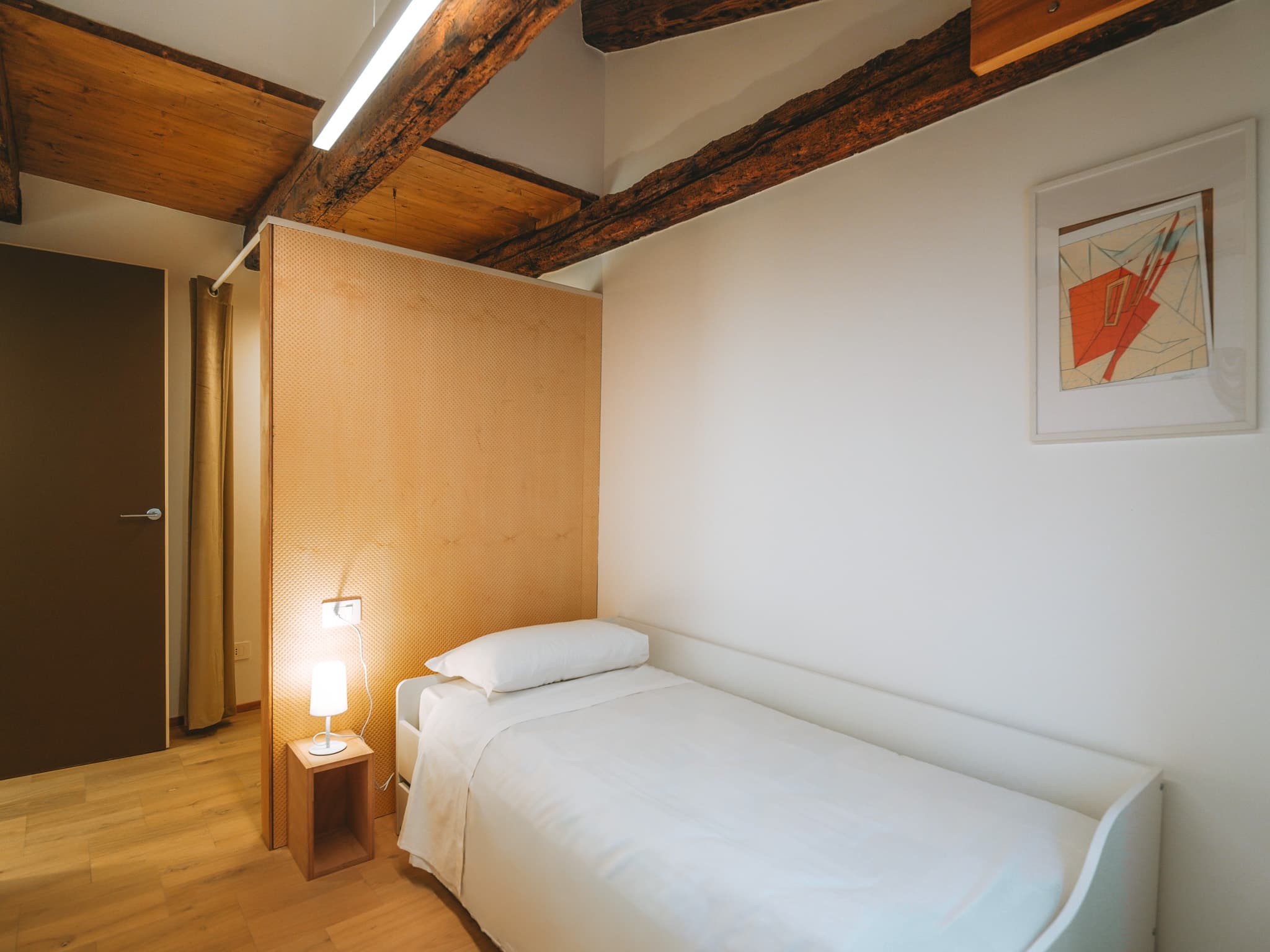 Camera con letto singolo bianco, pannello in legno chiaro, comodino con lampada e stampa astratta.