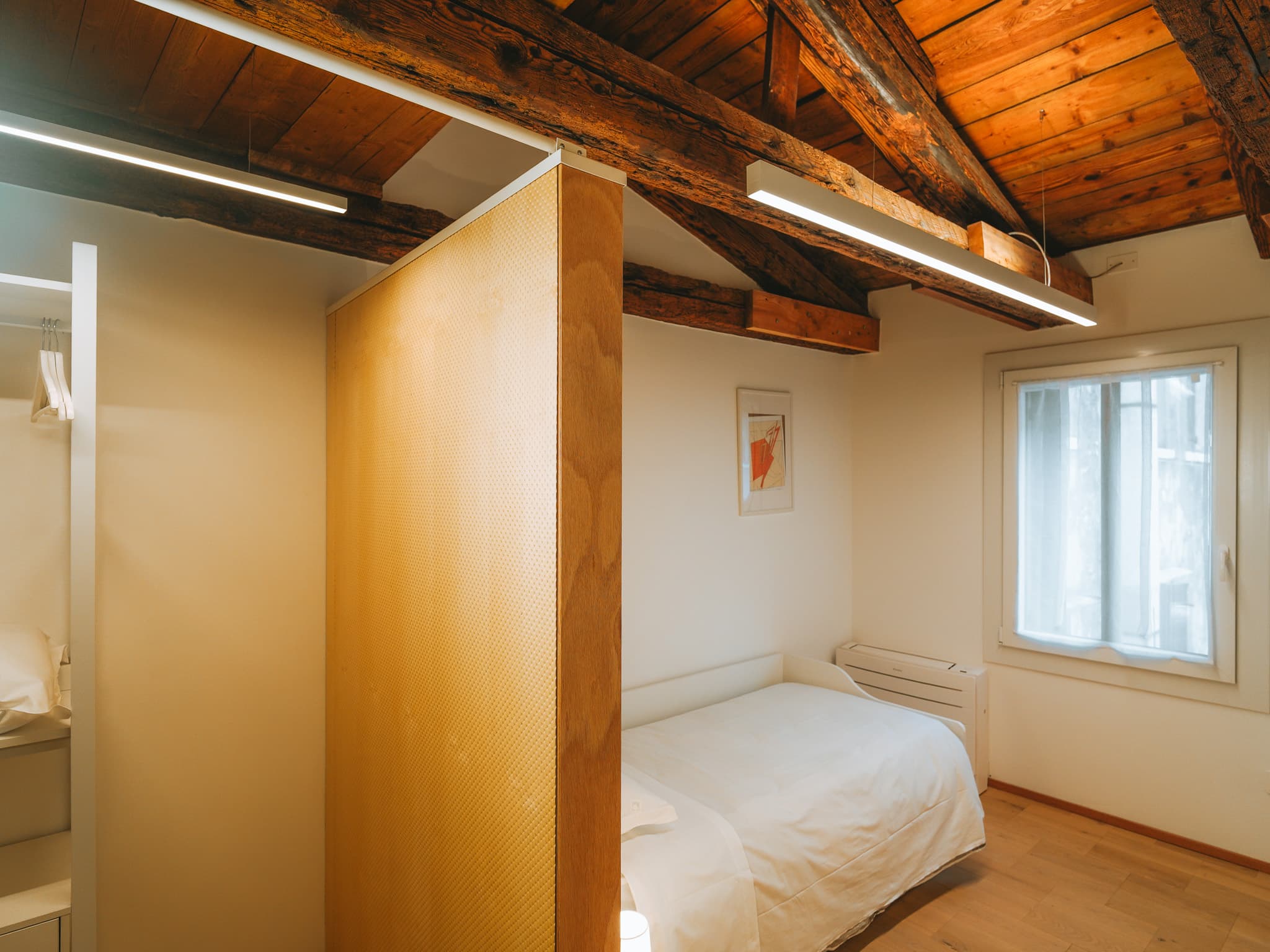 Letto singolo bianco accanto alla finestra, pannello divisorio in legno chiaro e travi a vista.