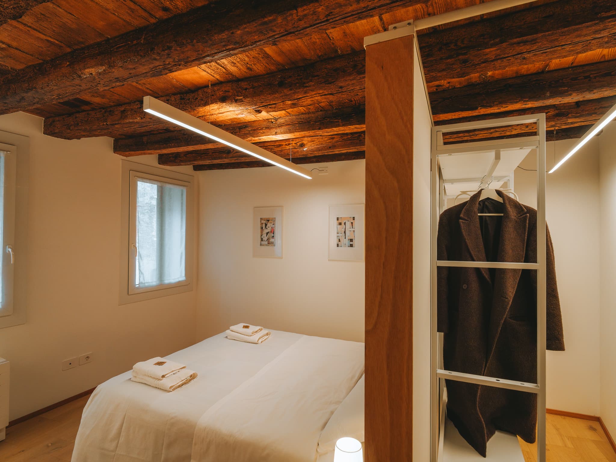 Camera con letto bianco, asciugamani piegati, due stampe incorniciate e cappotto appeso nell'armadio aperto.