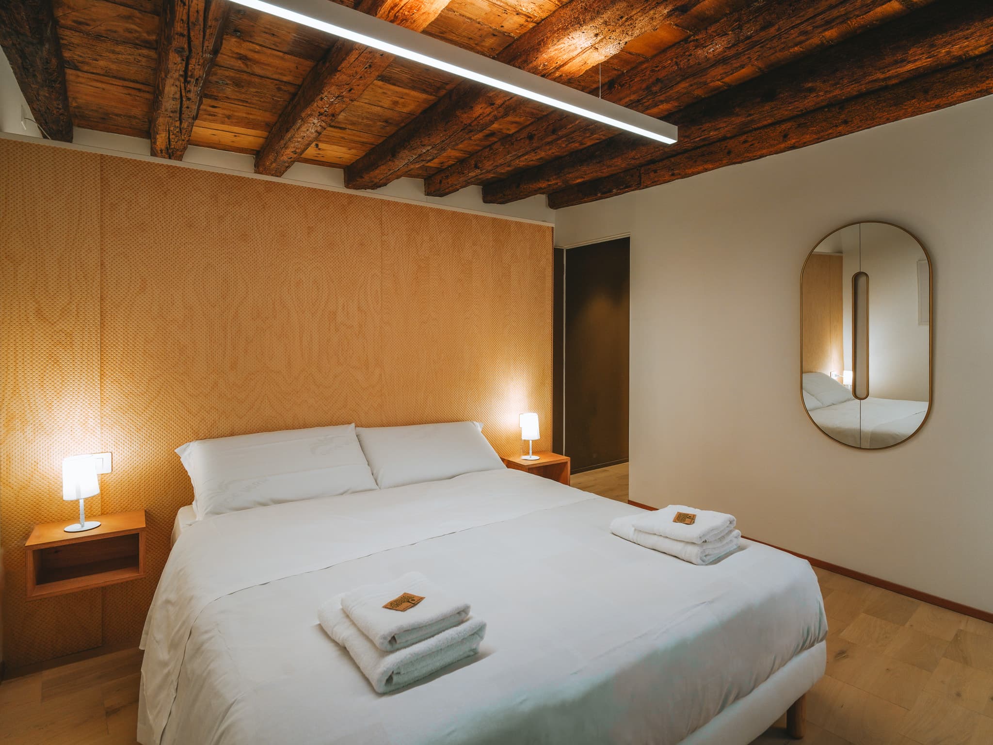 Letto matrimoniale con testiera in legno chiaro, comodini sospesi, lampade accese e specchio ovale.