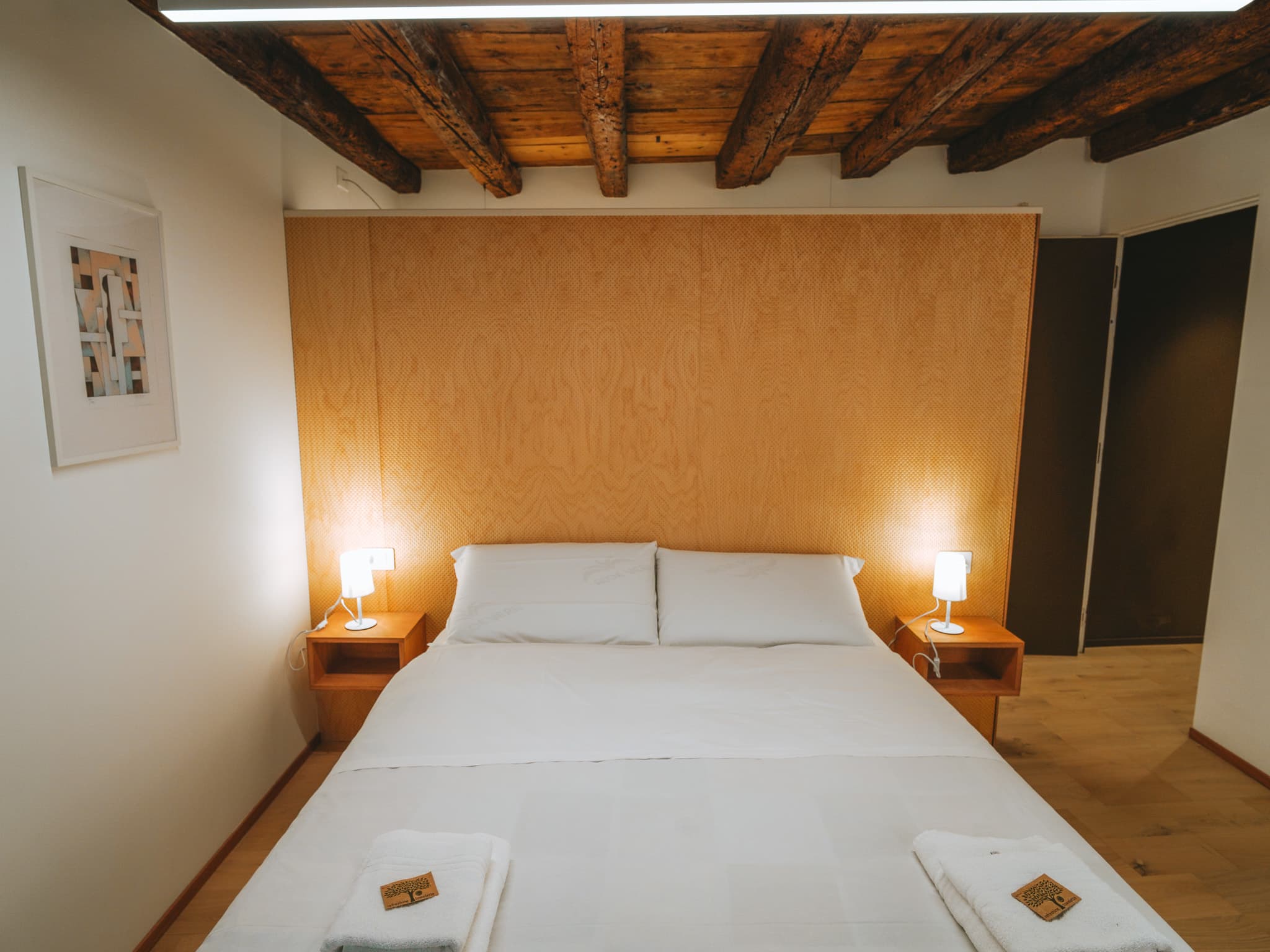 Letto matrimoniale visto frontalmente, testiera in legno traforato, due lampade accese e travi a vista.