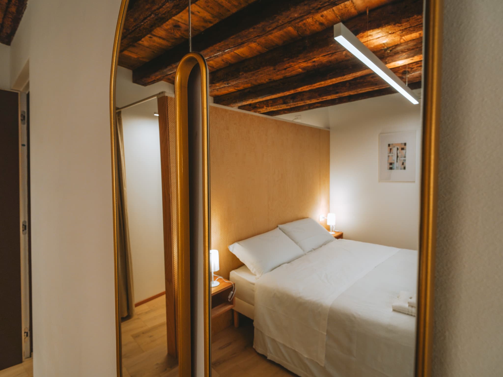 Specchio ovale con cornice dorata che riflette un letto matrimoniale, due comodini con lampade accese e il soffitto travato.