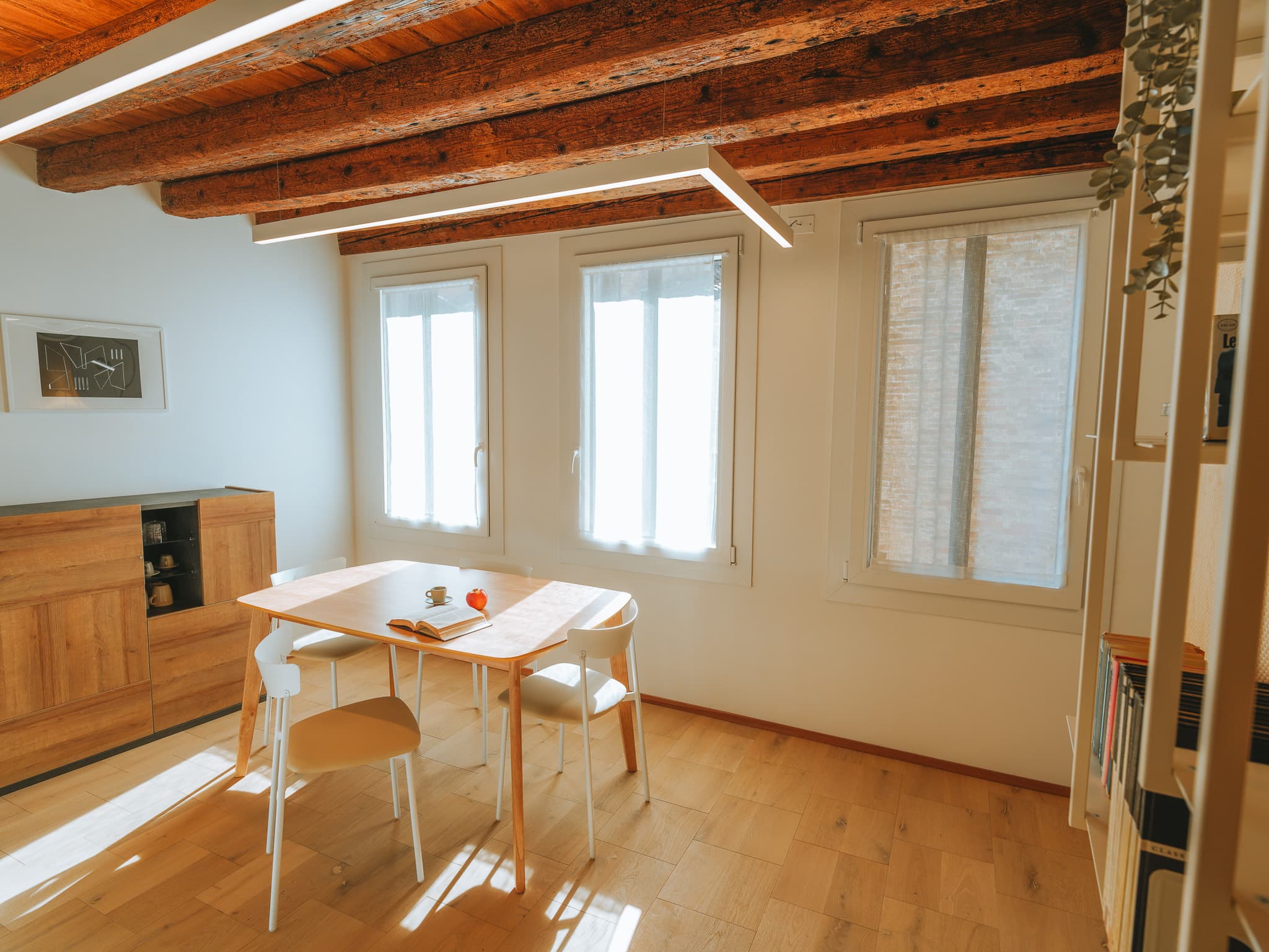 Tavolo in legno con quattro sedie bianche, credenza, tre finestre con tende, soffitto a travi.