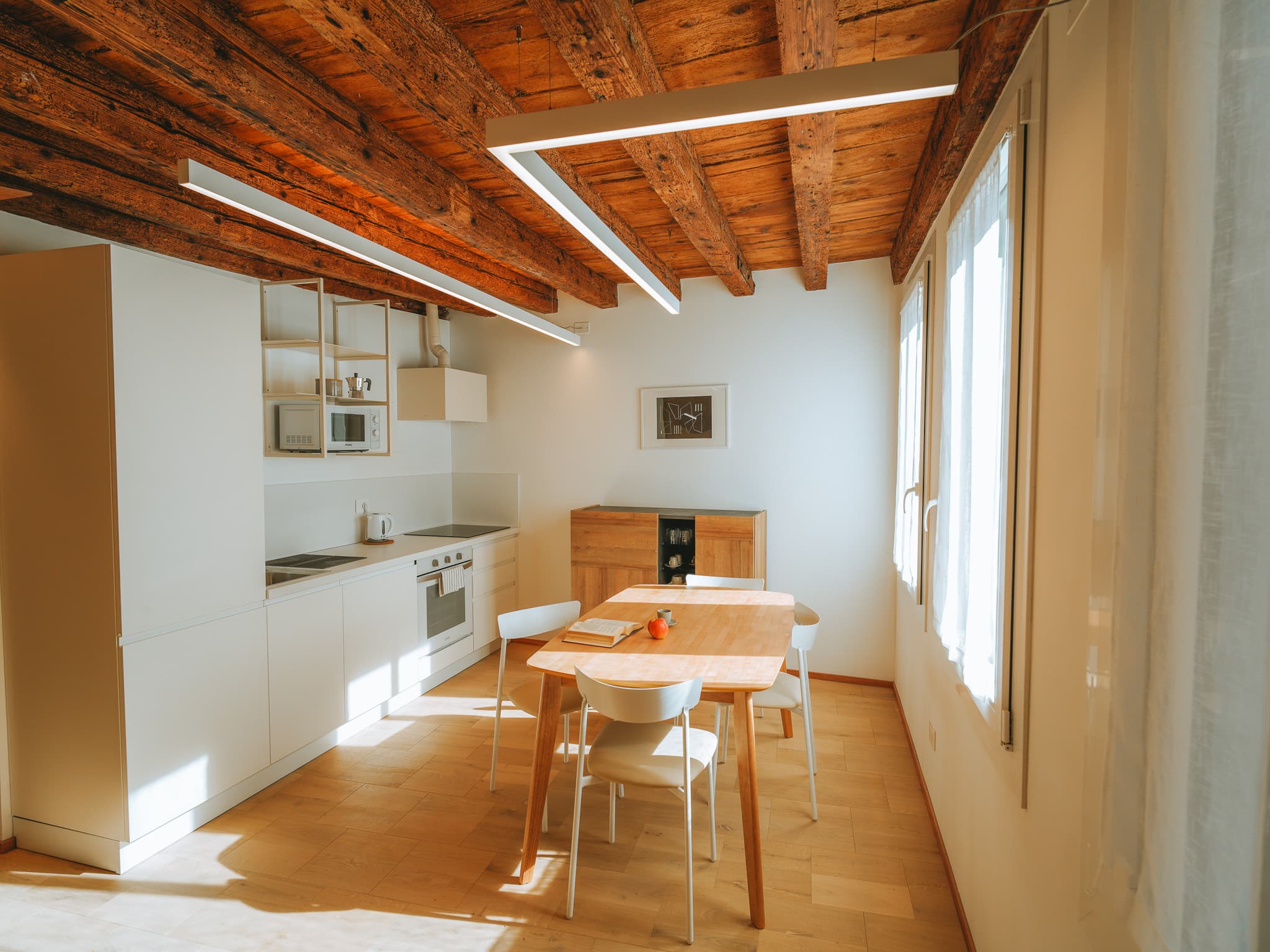 Cucina lineare bianca con forno e piano a induzione, mensole con moka e microonde, tavolo con sedie.
