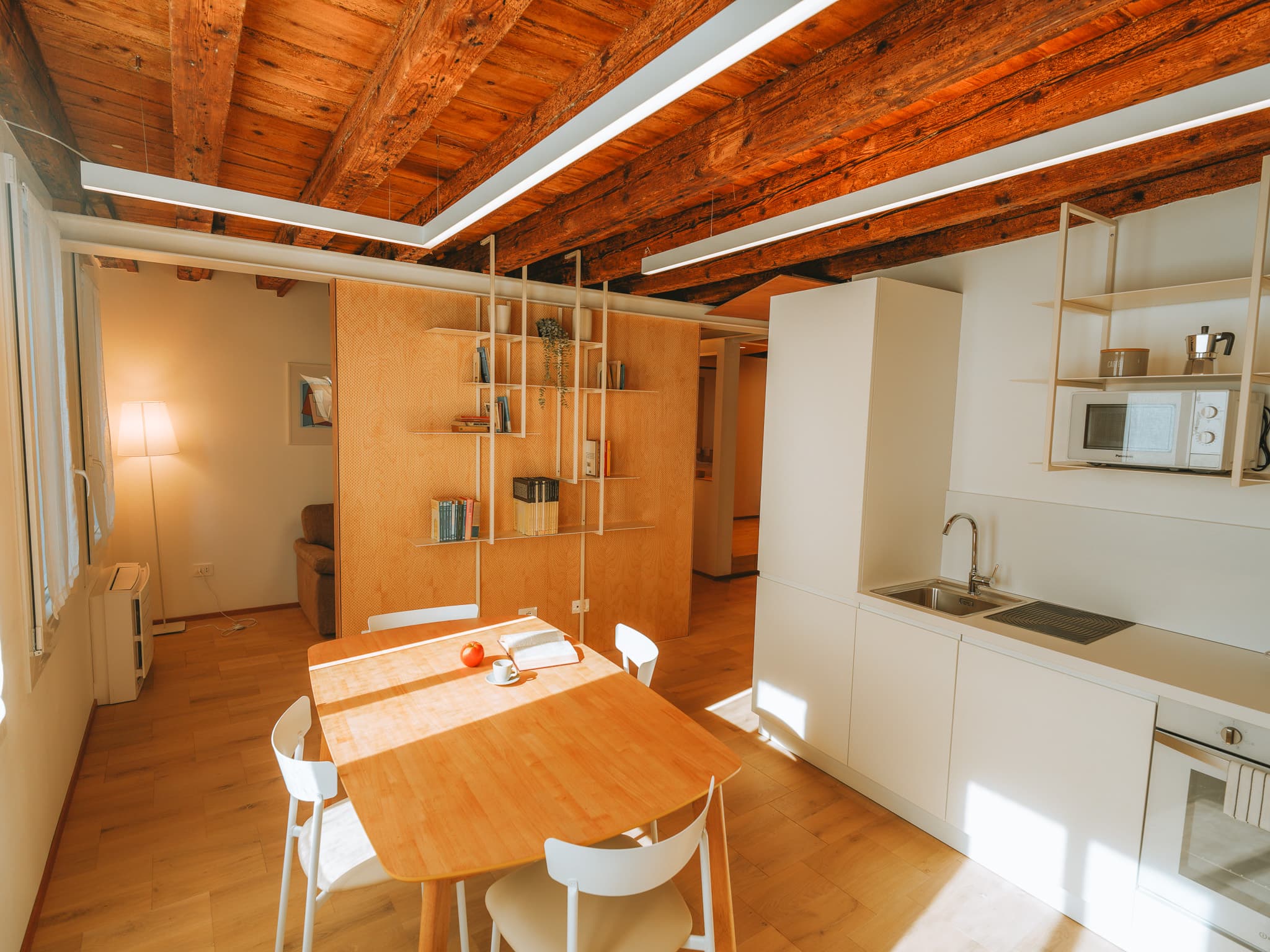 Cucina bianca con lavello e mensole, tavolo in legno con sedie bianche, parete divisoria in legno chiaro con libreria a mensole; sul fondo divano e lampada da terra, soffitto a travi.