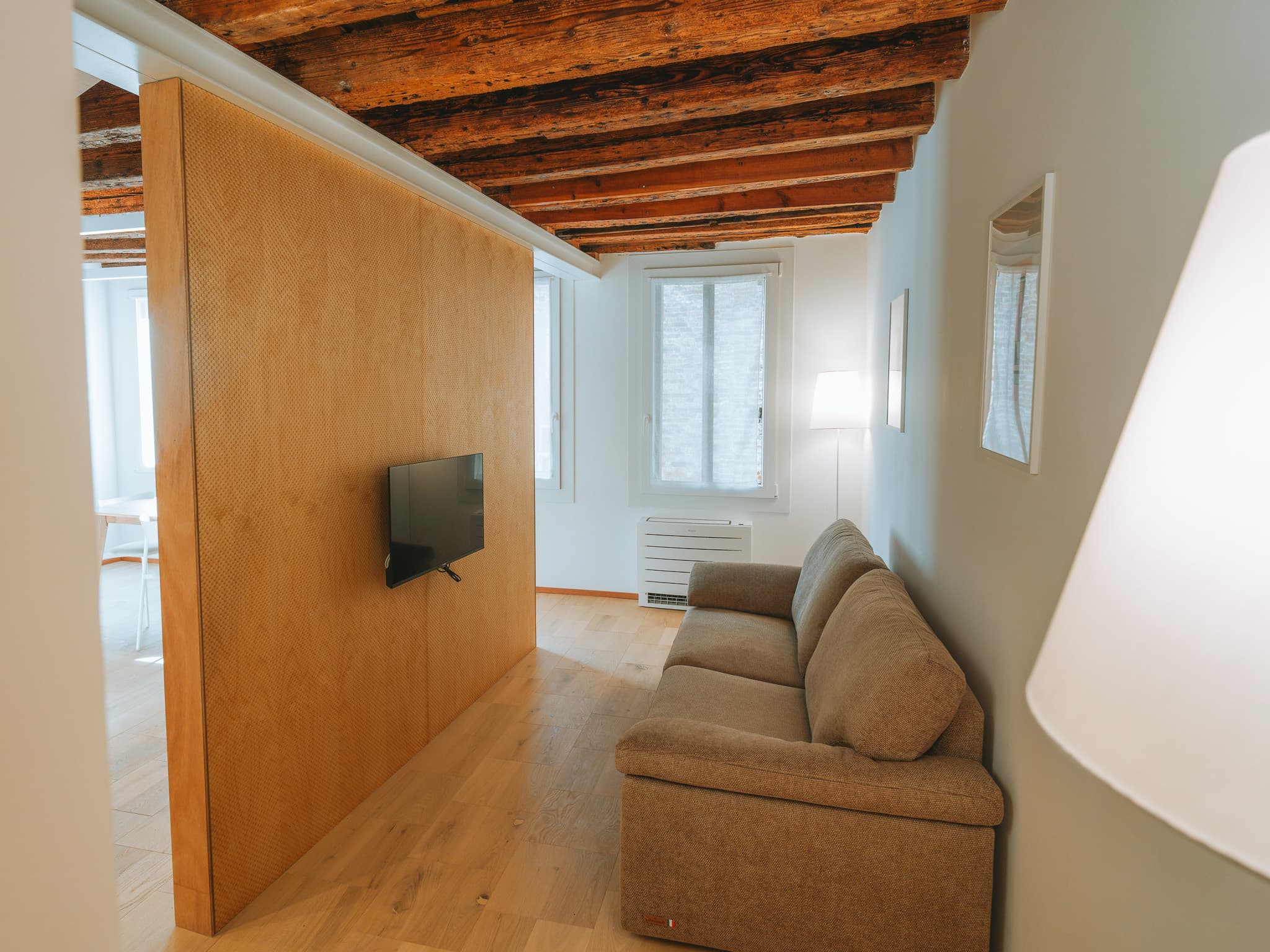Divano in tessuto marrone chiaro, televisore a parete su pannello in legno, lampada da terra accesa, due finestre con tende e due quadri incorniciati.