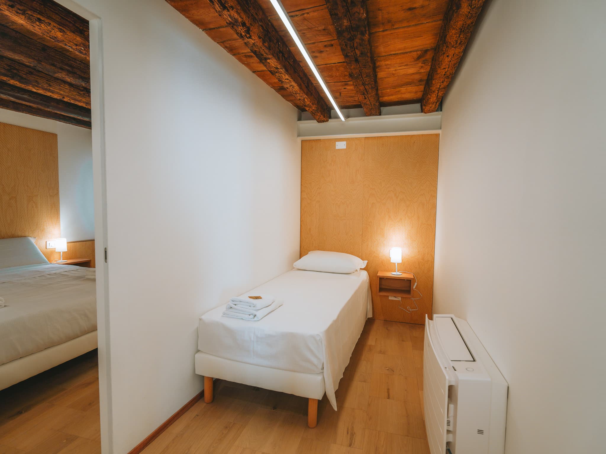 Letto singolo con asciugamani piegati, comodino con lampada accesa, porta aperta sul letto matrimoniale.