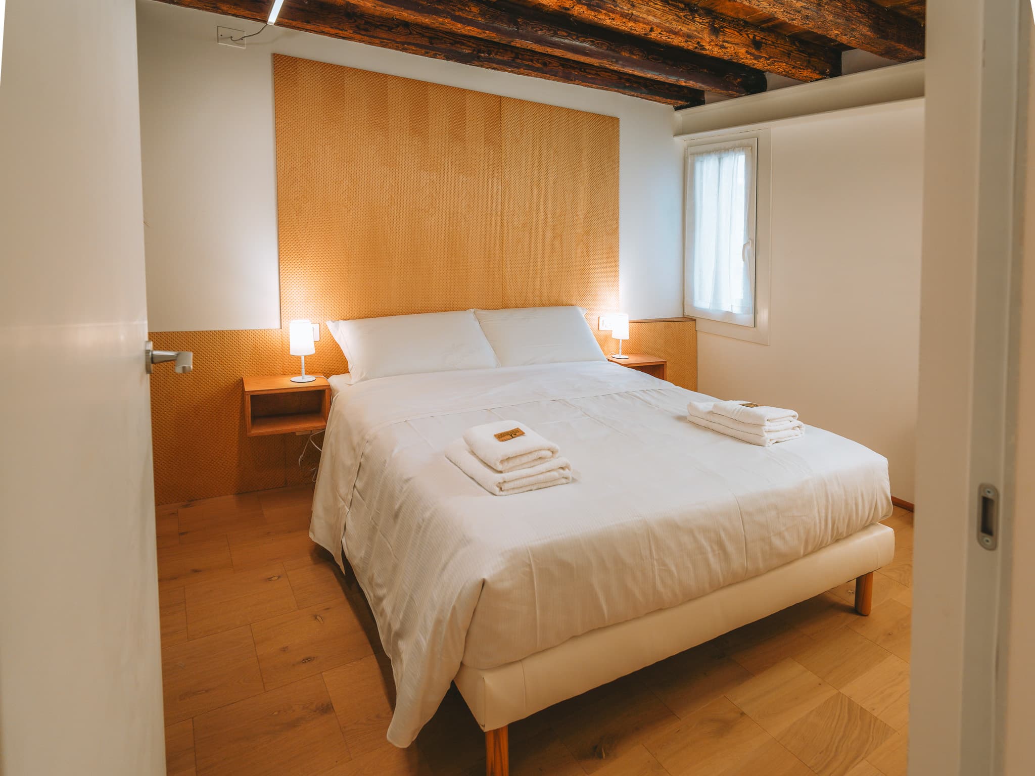 Letto matrimoniale con biancheria bianca e asciugamani piegati, due comodini sospesi con lampade accese, parete in legno chiaro dietro il letto, finestra con tenda.
