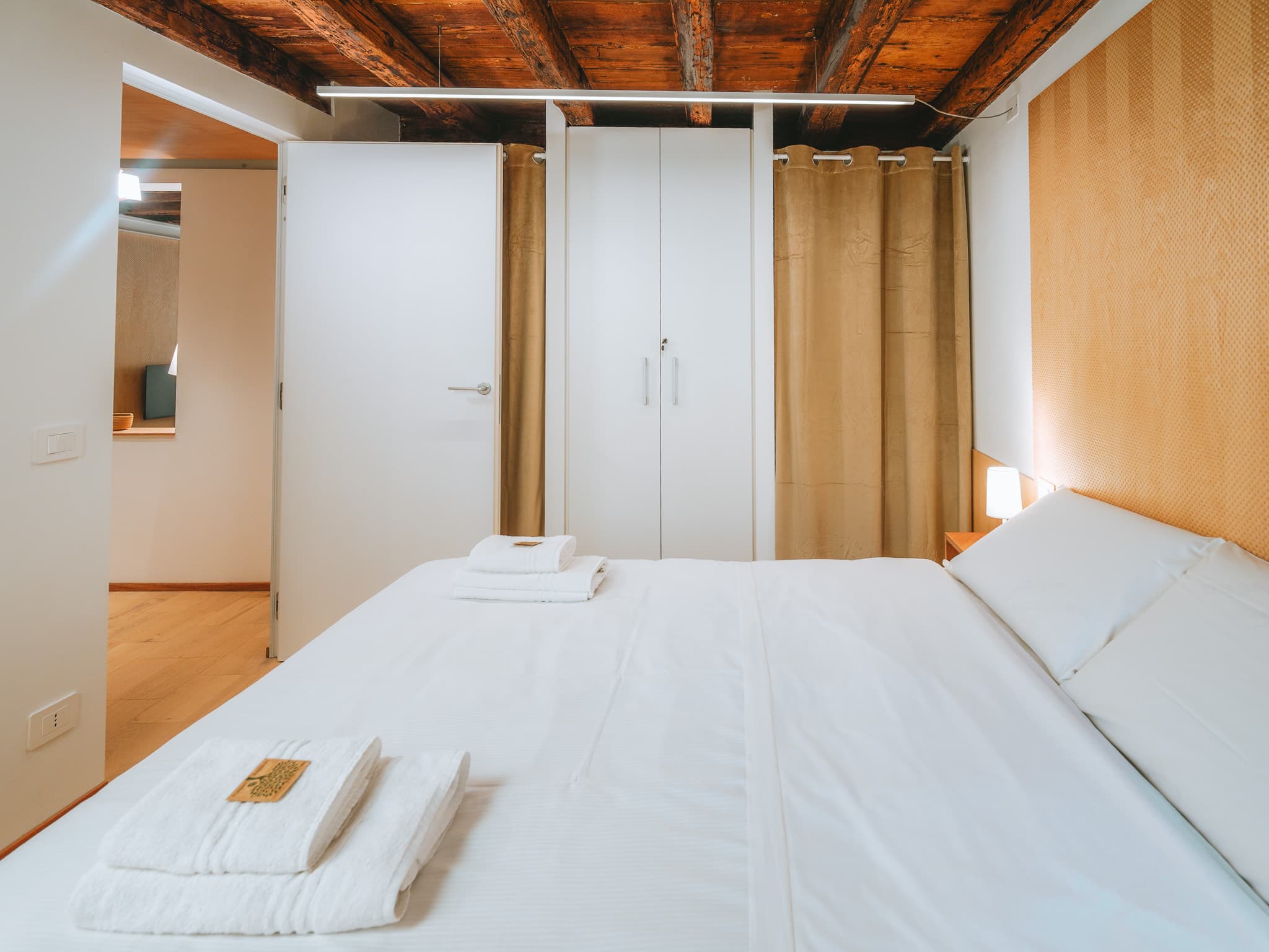 Letto matrimoniale con asciugamani piegati, armadio bianco a due ante, tende in velluto color oro, porta aperta sulla zona giorno con il televisore.