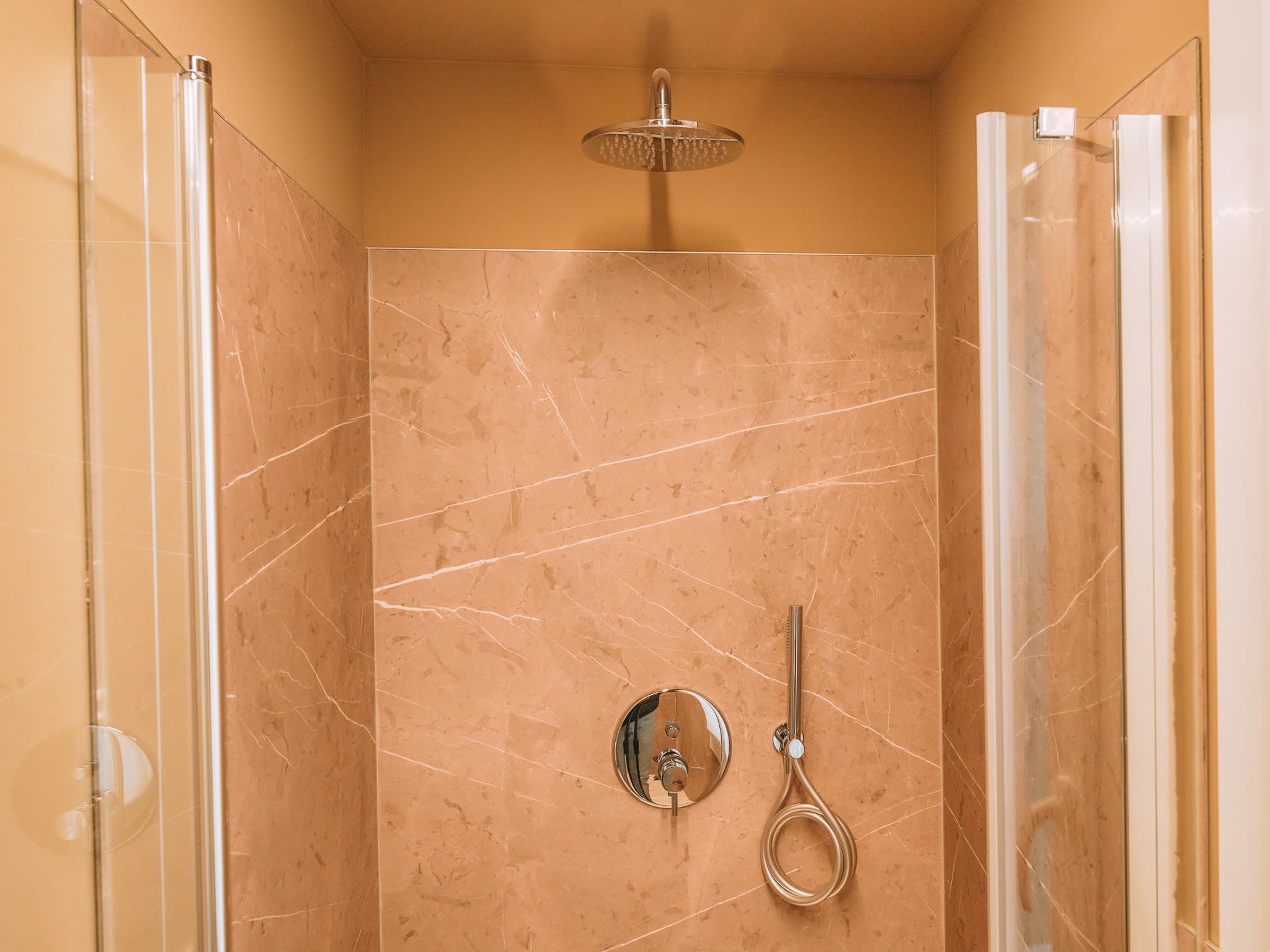 Doccia con soffione a pioggia, rivestimento in marmo beige venato, miscelatore e doccetta cromati.