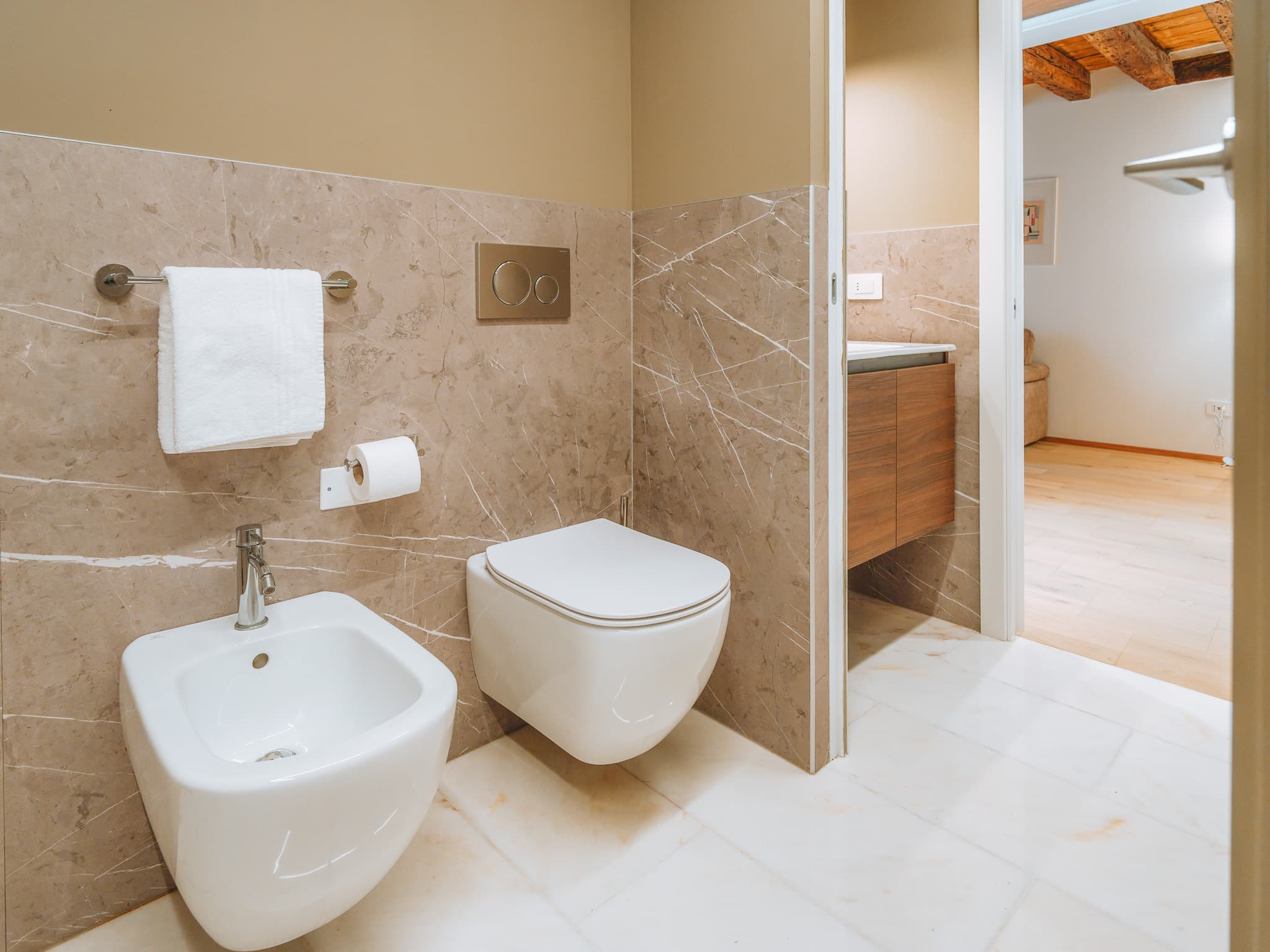Bidet e wc sospesi, rivestimento in marmo beige, asciugamano su portasalviette, porta aperta sul soggiorno.