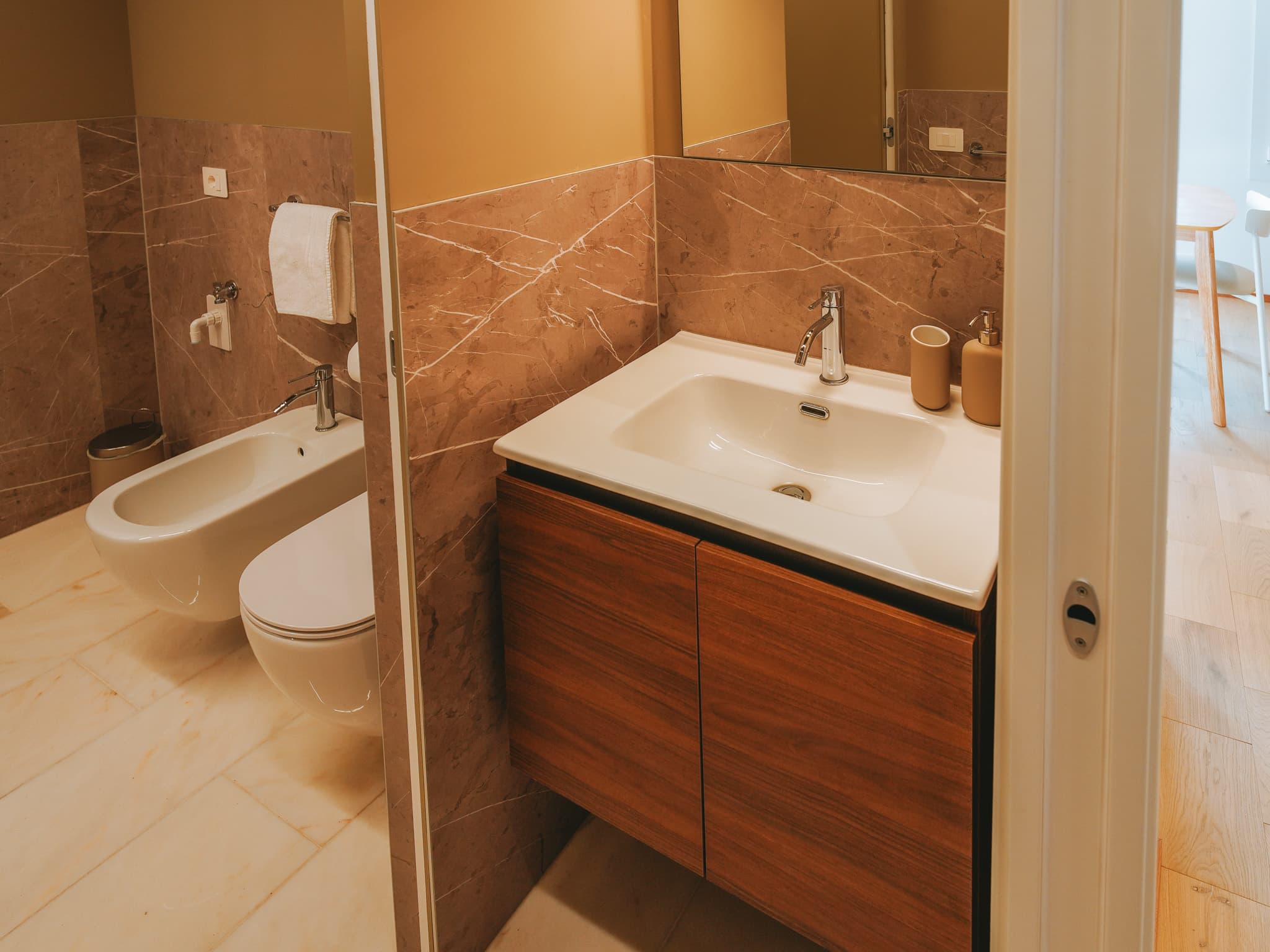 Lavabo bianco su mobile in legno, specchio, dosatore e bicchiere, bidet e wc oltre la parete.