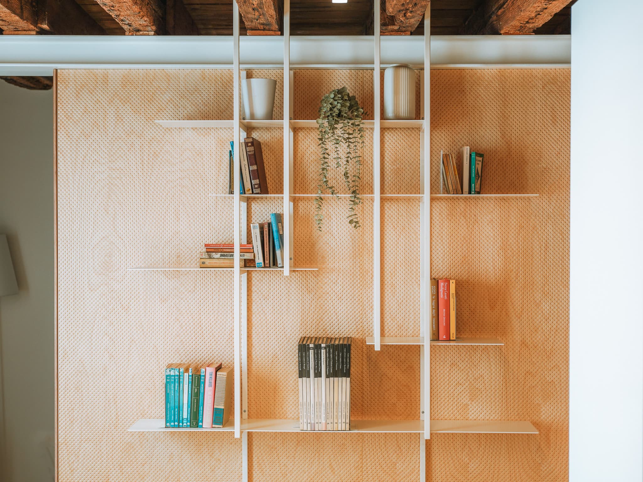 Libreria con mensole bianche su parete in legno traforato, libri, vasi in ceramica e pianta ricadente.