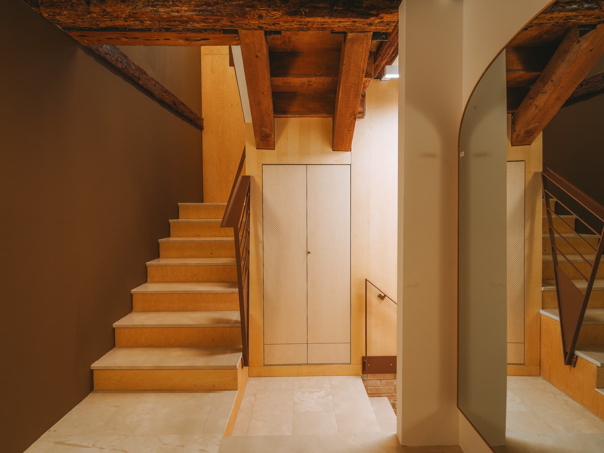 Scala in pietra chiara con corrimano in metallo brunito, travi di legno a vista e specchio arcuato.