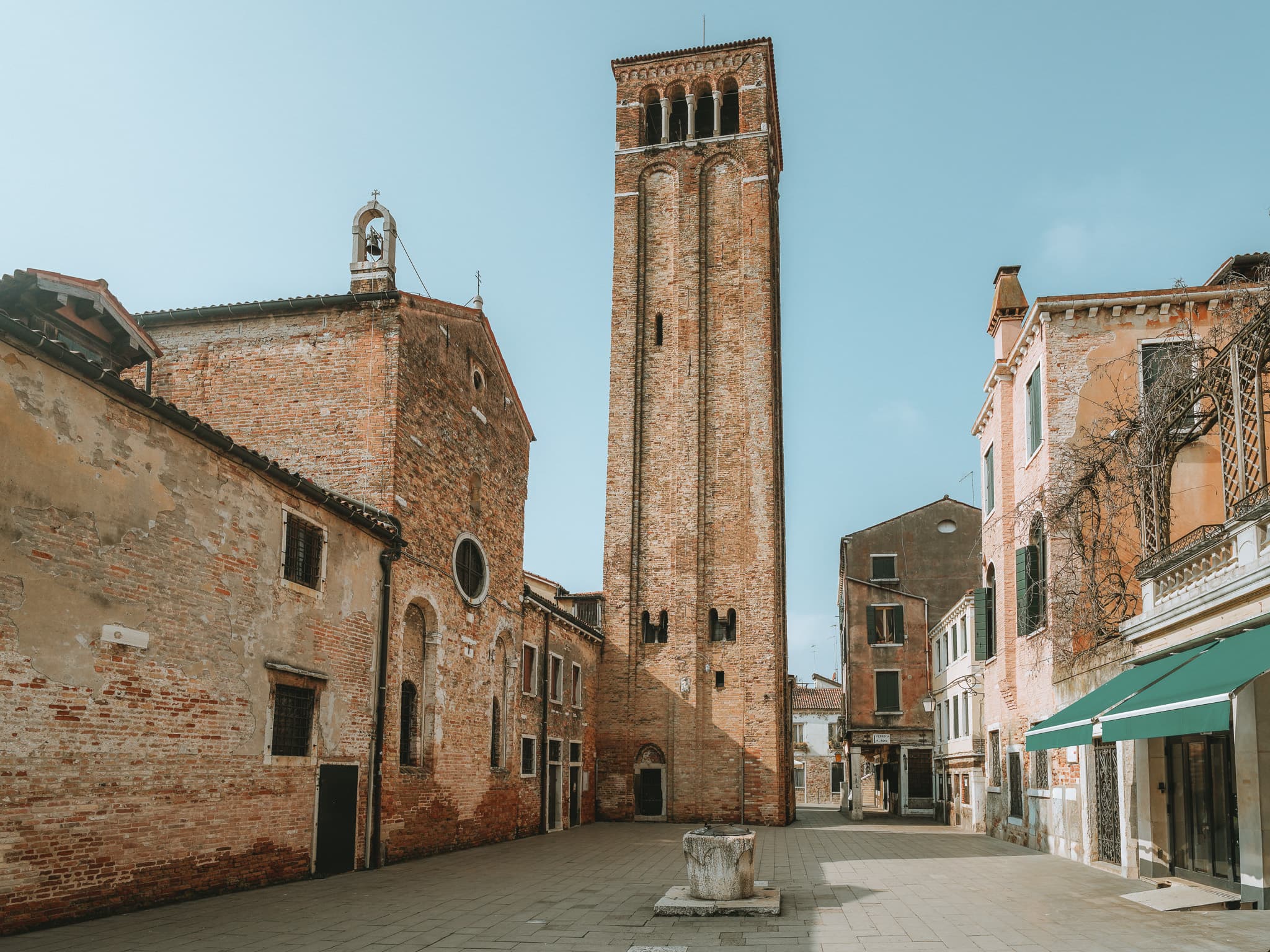 Campo pavimentato con chiesa in mattoni, alto campanile e vera da pozzo in pietra al centro.